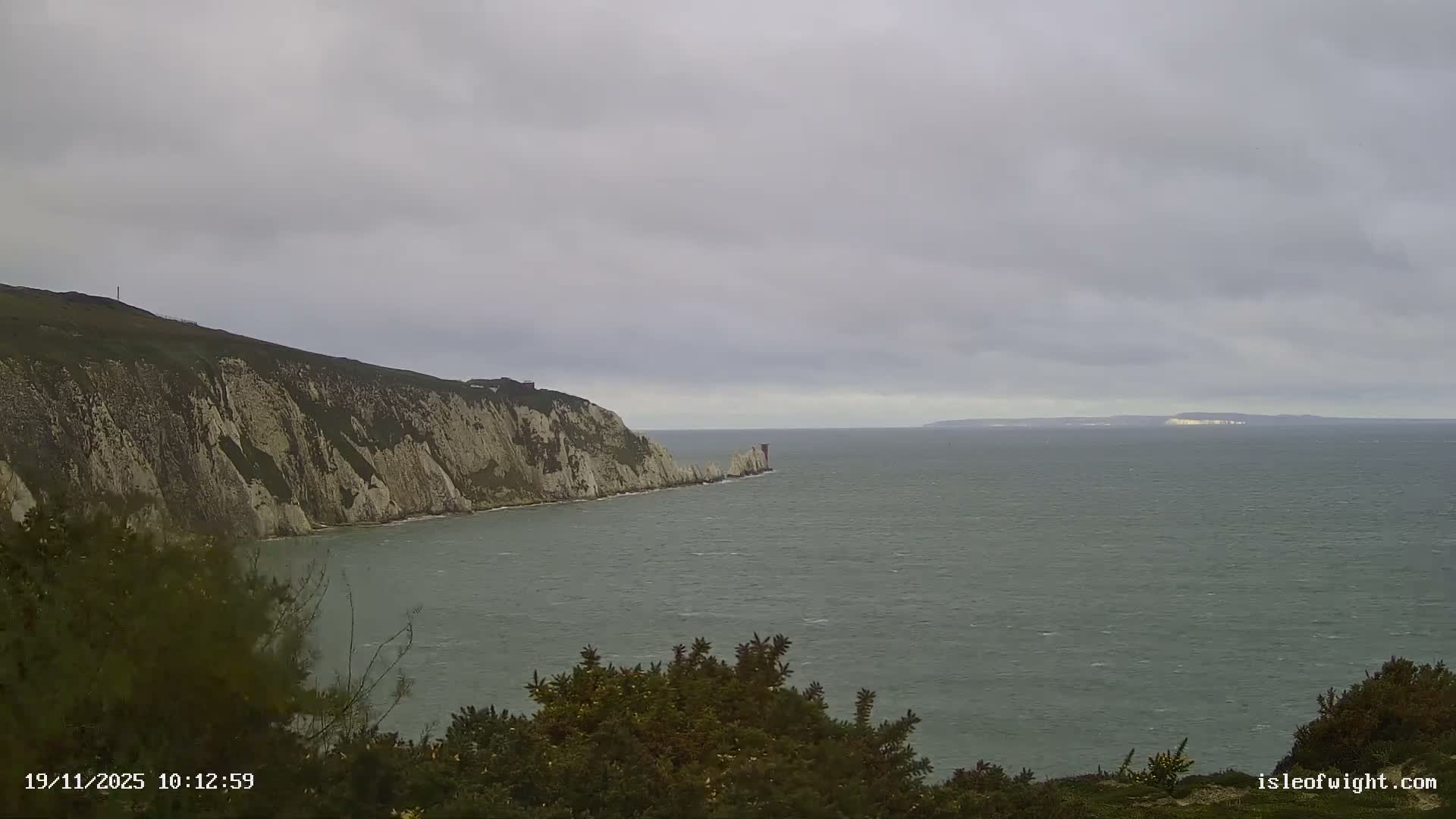 An outdoor scene features rugged white cliffs meeting a choppy gray sea, with a distinctive lighthouse on a rock formation and a distant landmass visible under a heavily overcast sky.