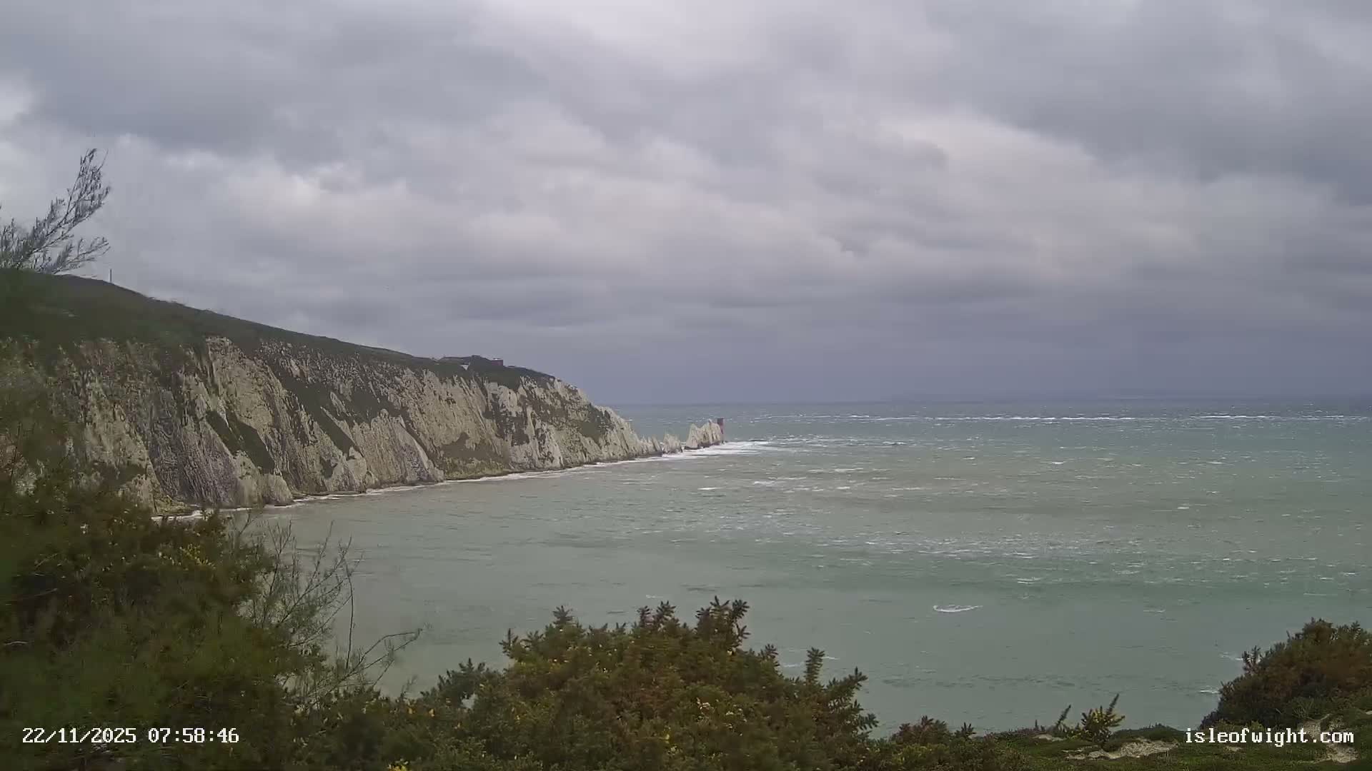 A rugged coastline features prominent white cliffs and rocky sea stacks jutting into a turbulent, grey-green sea under a heavily overcast and gloomy sky.