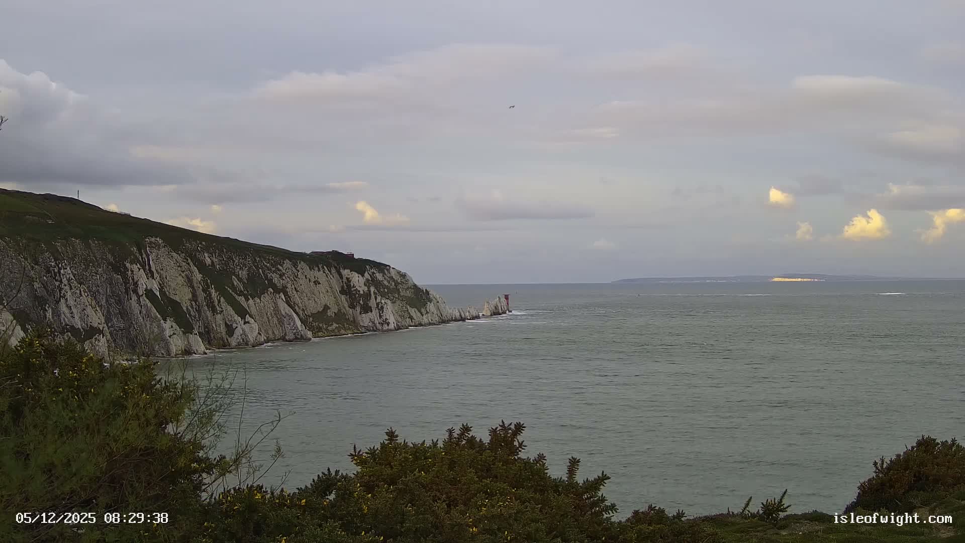 A coastal landscape on a mostly cloudy morning features prominent white chalk cliffs and The Needles lighthouse reaching into the choppy sea, with distant land glowing faintly on the horizon.