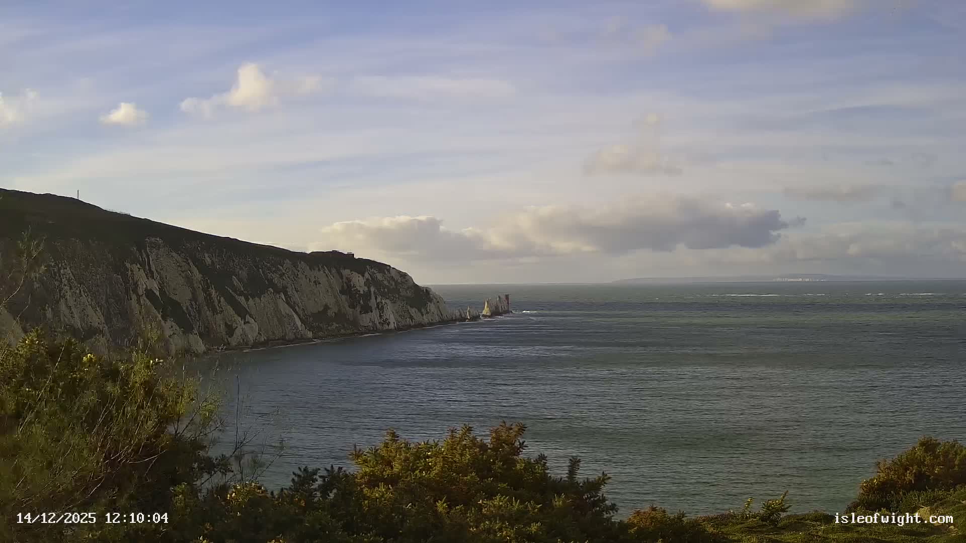 A coastal landscape on a mostly cloudy morning features prominent white chalk cliffs and The Needles lighthouse reaching into the choppy sea, with distant land glowing faintly on the horizon.