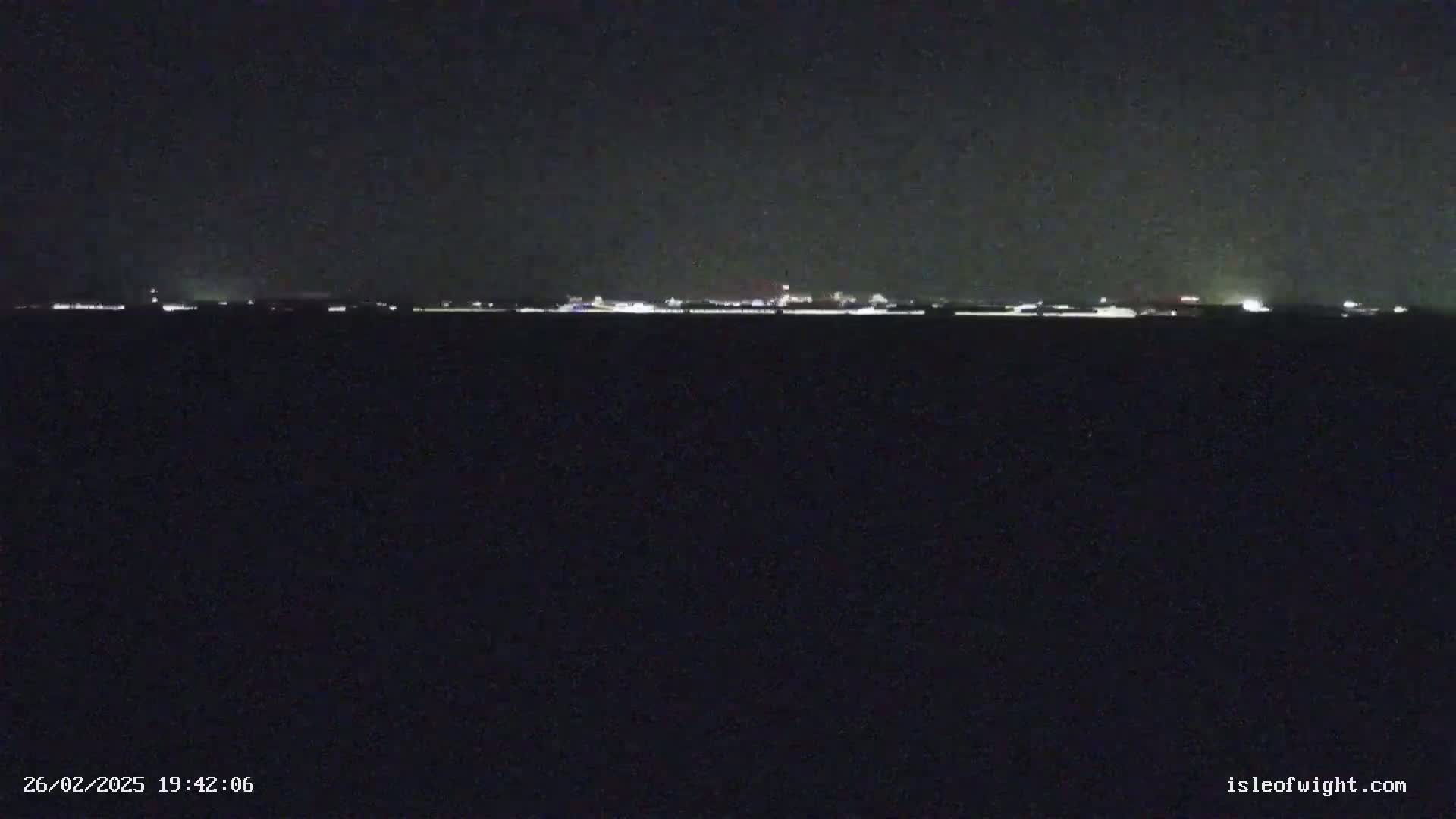 A dark, nighttime scene shows a distant line of lights across a dark body of water under a clear, dark sky.