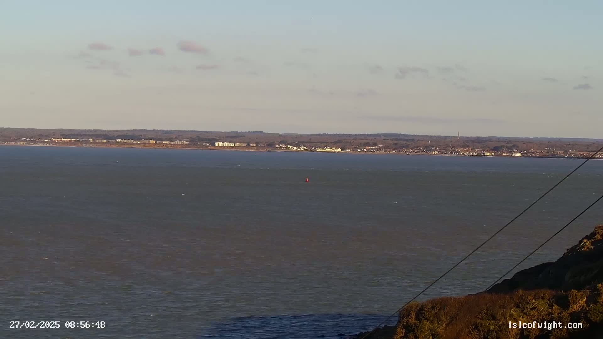 A calm, gray body of water stretches to a distant shoreline under a mostly clear sky, with a small portion of land visible in the foreground.