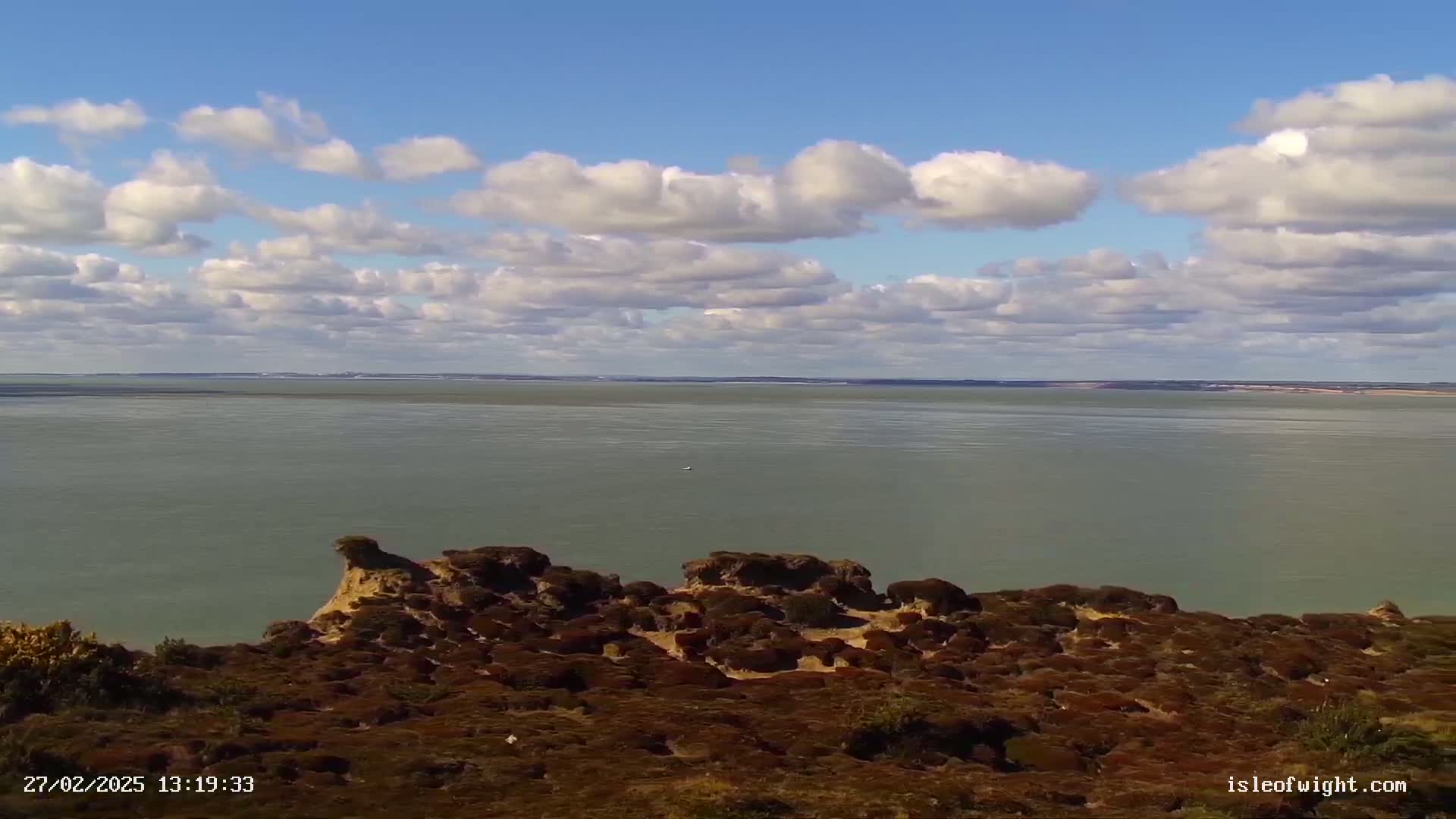A tranquil expanse of water stretches to the horizon under a partly cloudy sky, viewed from a vantage point overlooking low-lying, dark-brown vegetation and rocky formations.