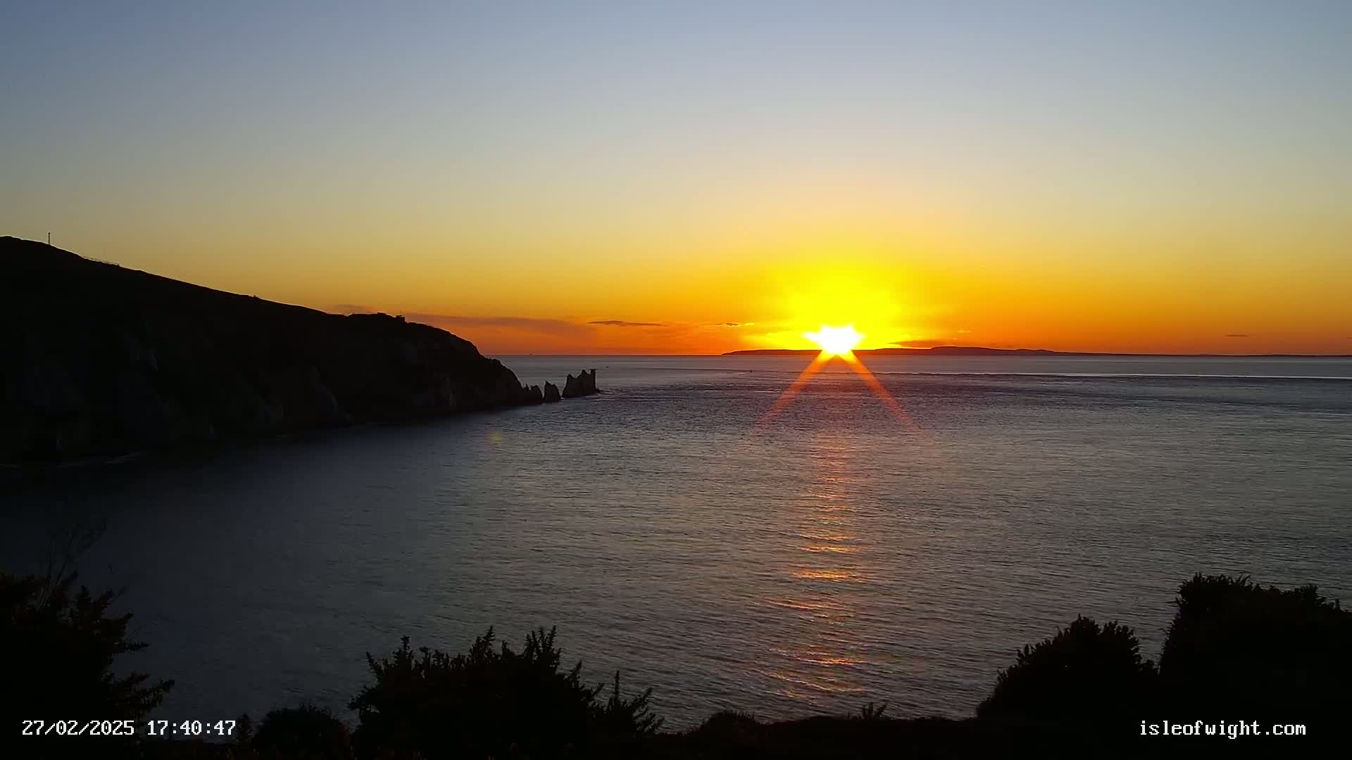 A vibrant sunset over a calm sea, casting a golden glow on the water, with a dark, rocky coastline in the foreground.