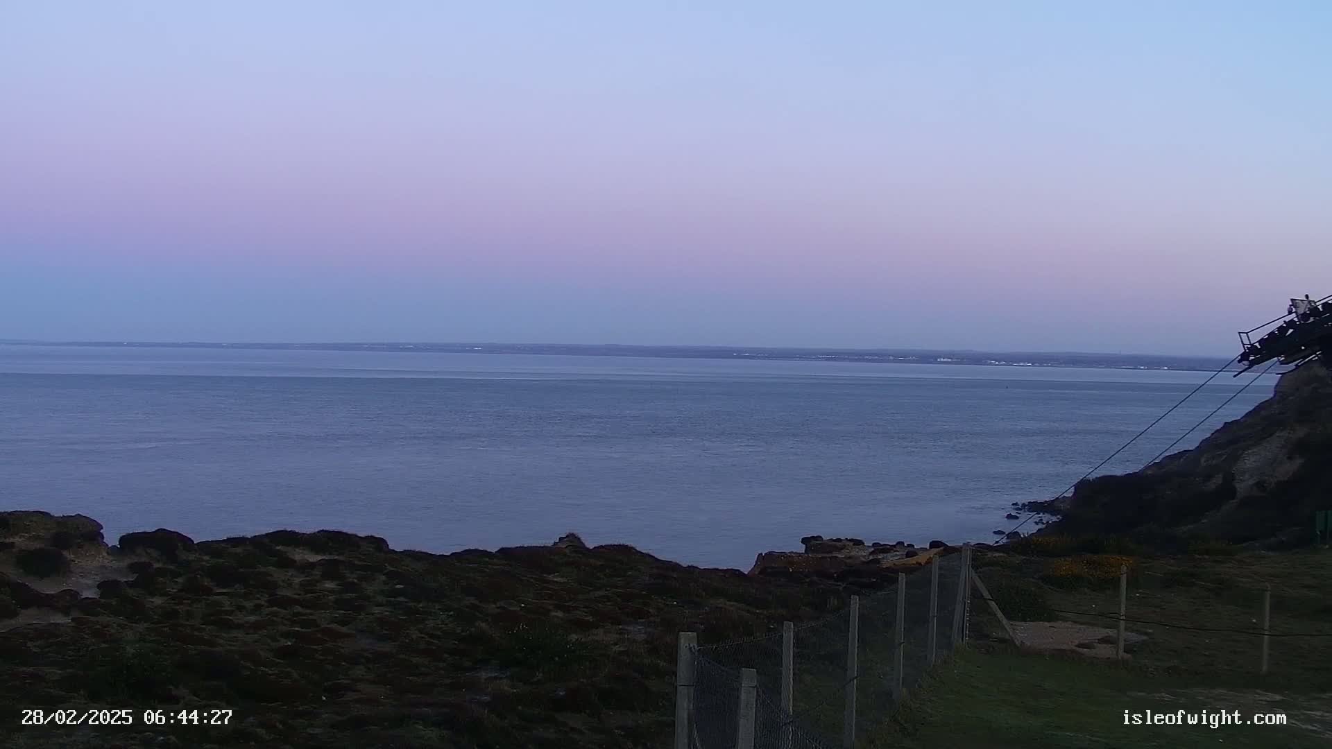 A calm sea stretches to a pale lavender horizon under a twilight sky, viewed from a clifftop with a chain-link fence in the foreground.
