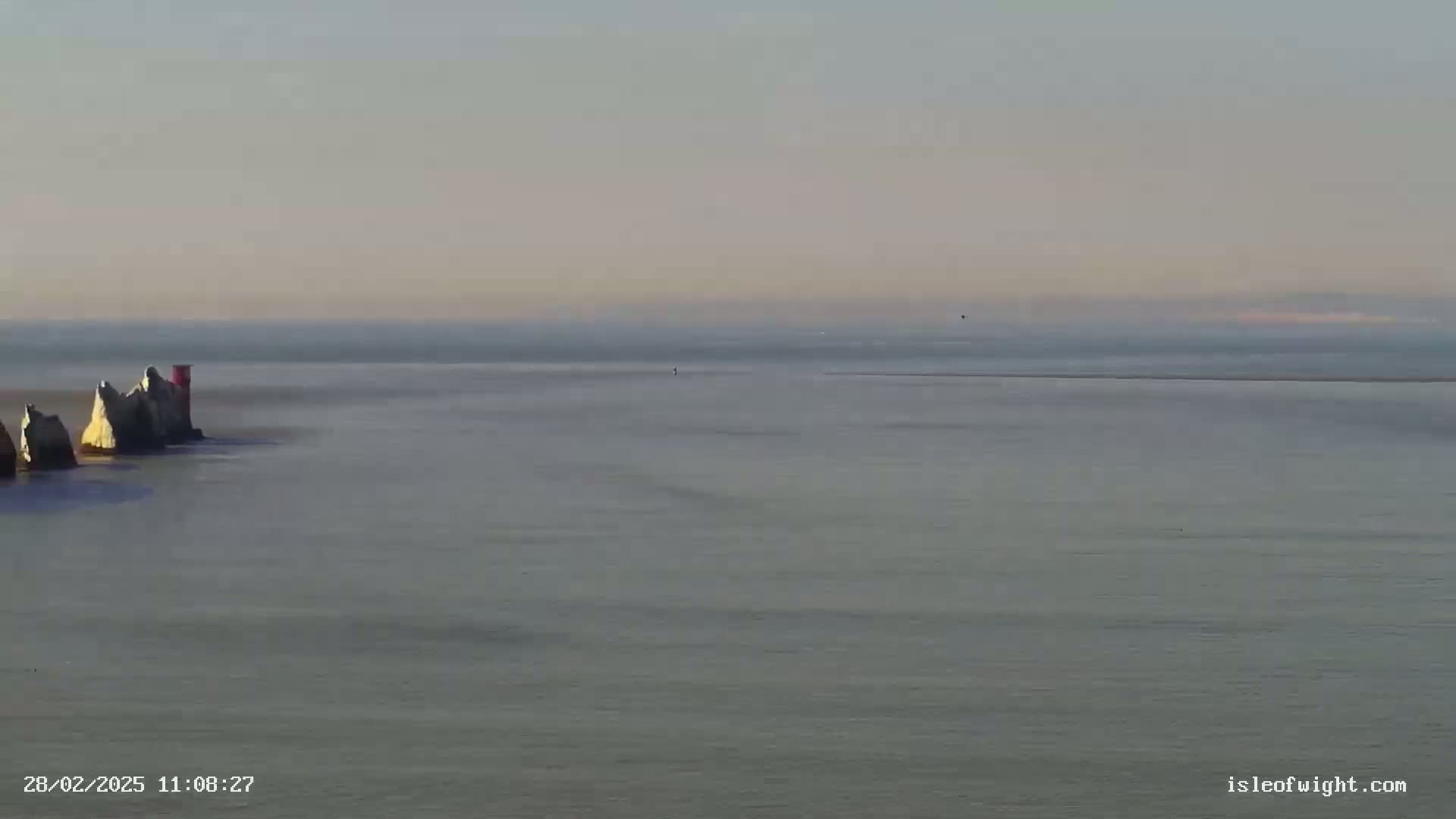 A calm, hazy sea stretches to the horizon, featuring a group of white rock formations with a small red structure on one, under a pale sky.
