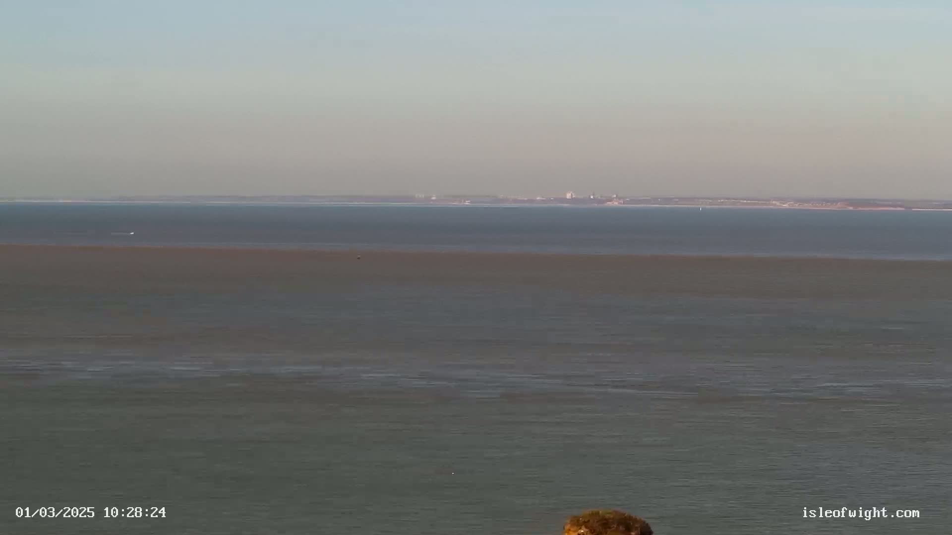 A calm, wide expanse of water shows a subtle color difference in its layers, with a distant shoreline visible on the horizon under a clear sky.