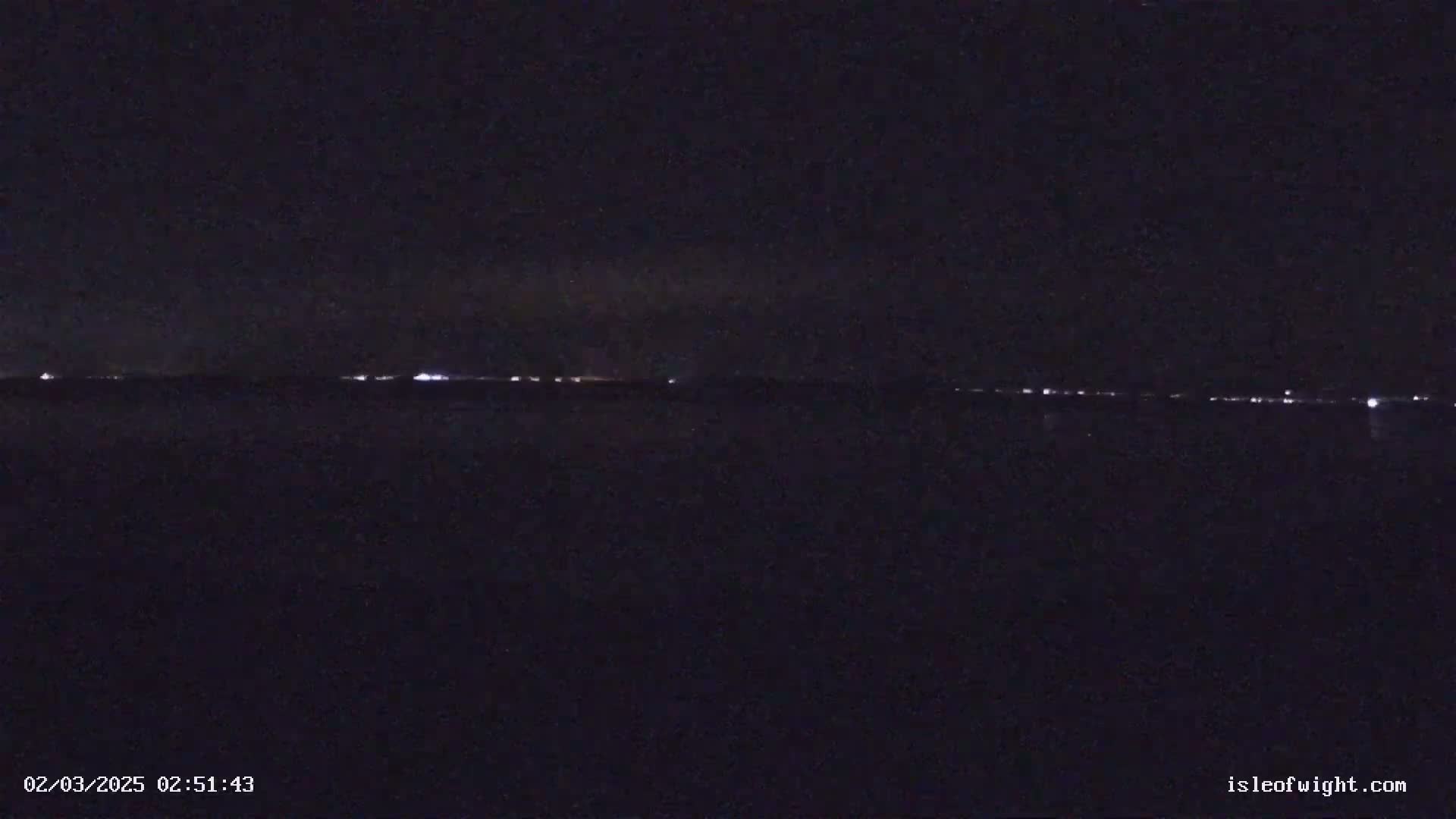 A dark, nighttime scene shows a distant line of lights across a body of water under a clear, dark sky.