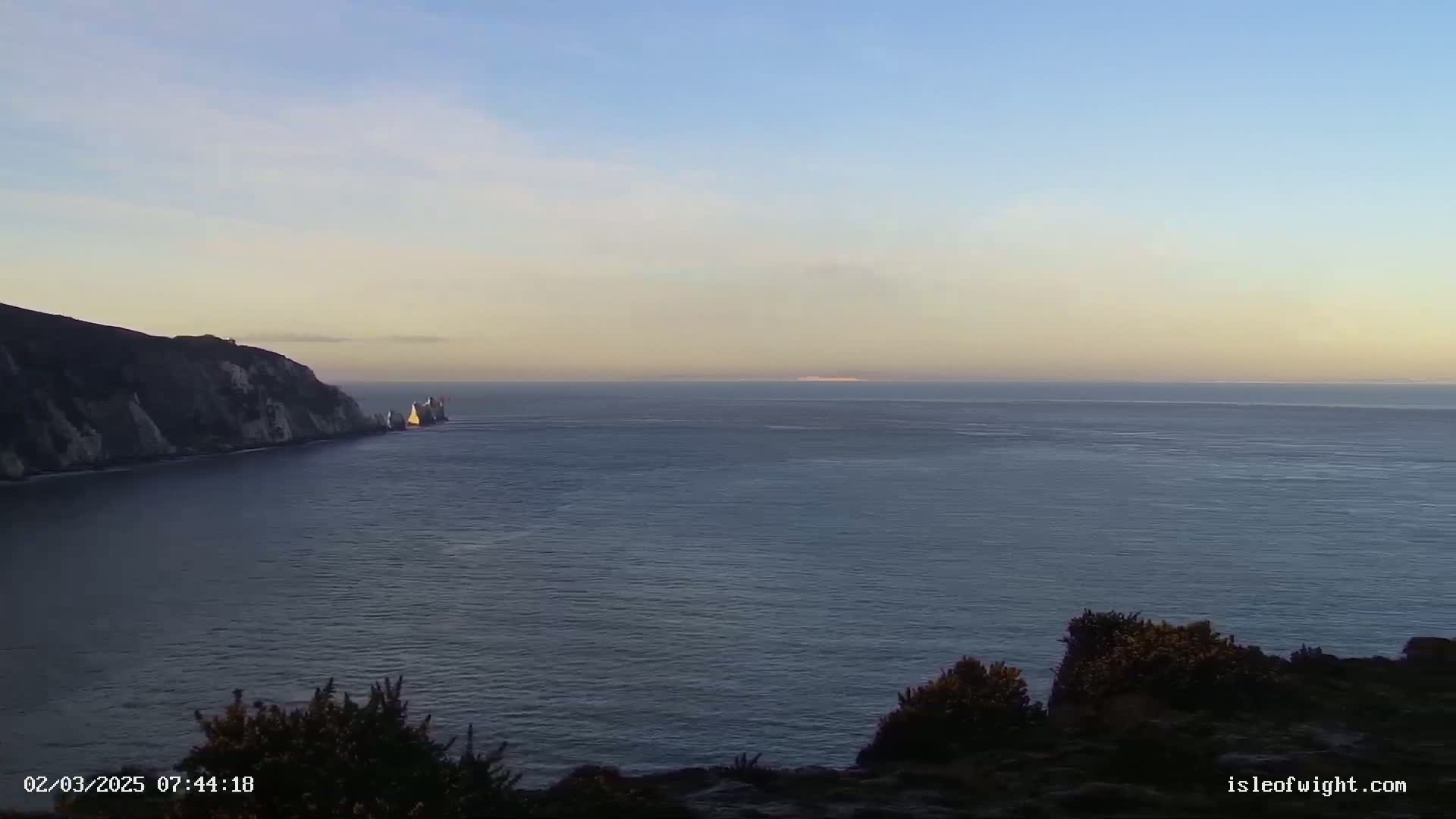 A calm, twilight sea stretches to a distant horizon, where a faint landmass is visible; a dark cliff and a group of light-colored rocks jut out from the sea on the left.