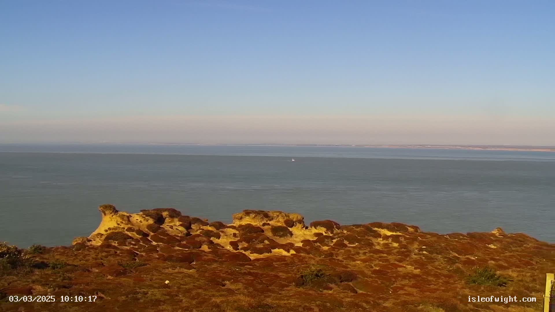 A rocky, reddish-brown hillside covered in low vegetation overlooks a calm, gray sea under a clear blue sky.