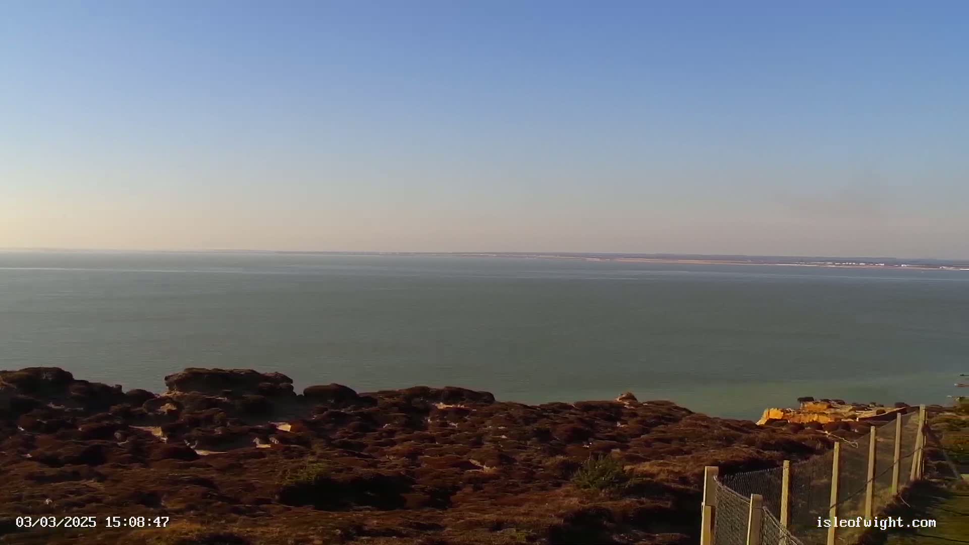 A calm, expansive body of water stretches to a distant landmass under a clear, bright sky, viewed from a rocky, vegetated clifftop with a fence.