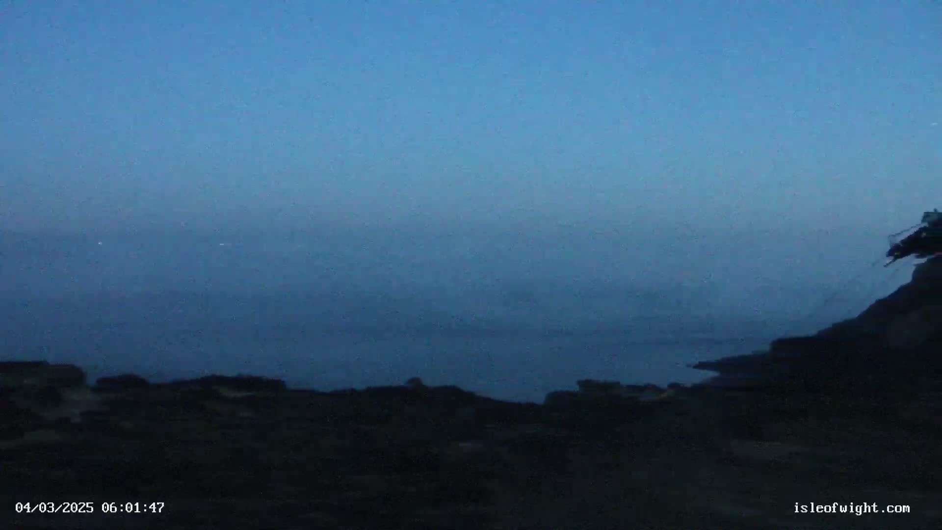 A dark, foggy coastal scene is visible at twilight, with a portion of land in the foreground and a body of water extending to a hazy horizon under a dusky blue sky.