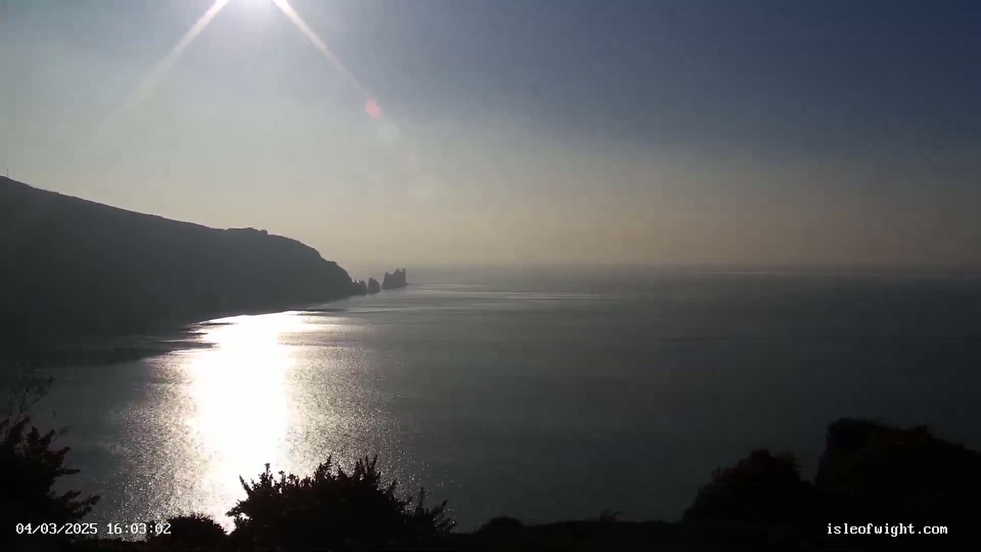 A bright sun shines over a calm sea reflecting its light, with a dark cliff in the foreground and another visible in the distance.