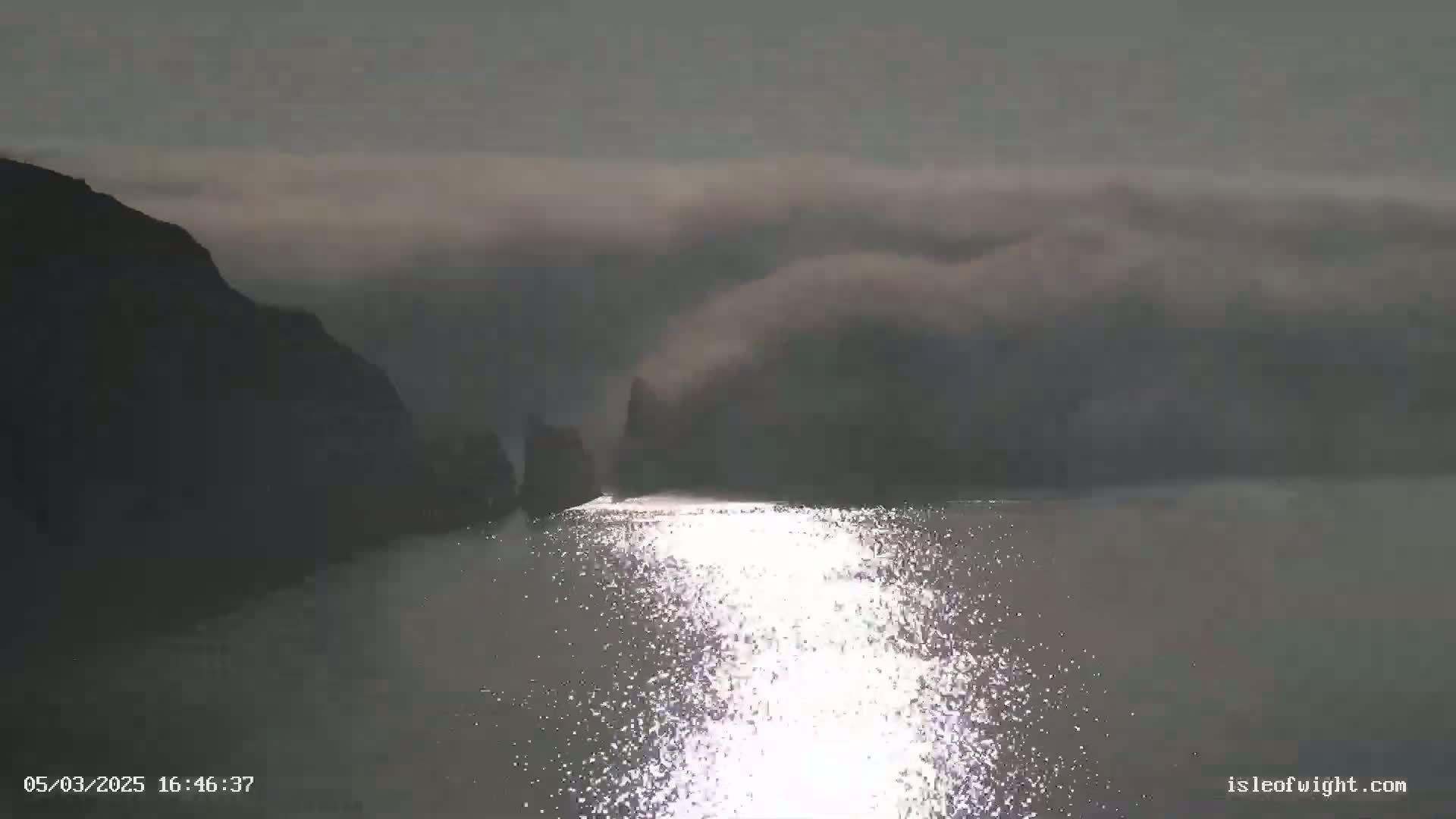 Dark, rocky cliffs are partially obscured by fog over a calm body of water reflecting sunlight.