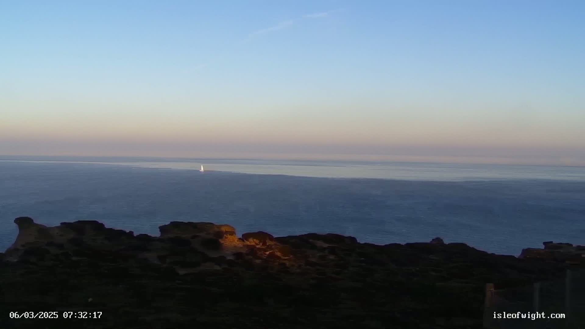 A sailboat is visible on a calm, twilight sea from a dark, rocky coastal vantage point.