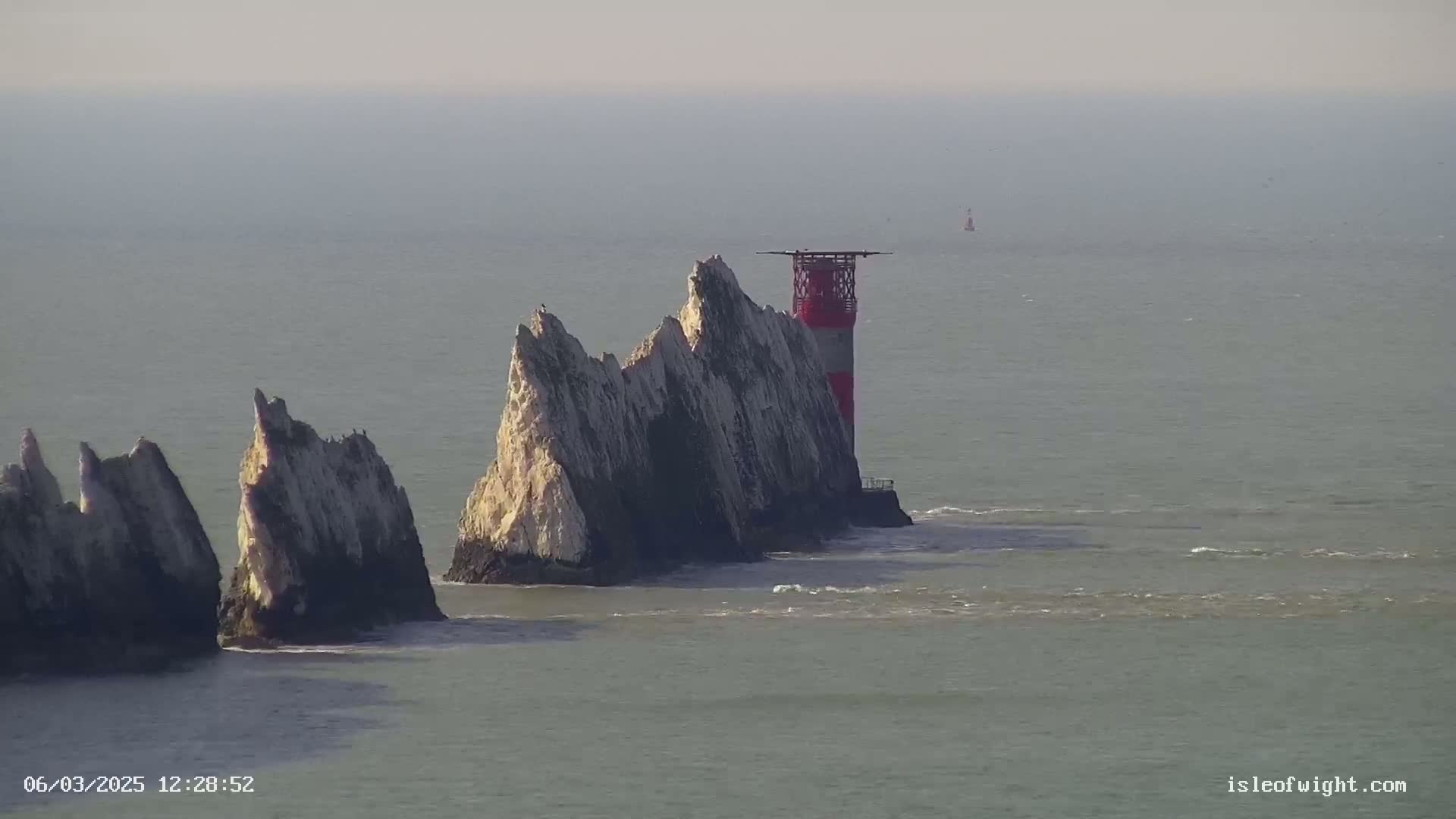 A red and white lighthouse stands atop a rocky outcrop in the sea on a hazy day.