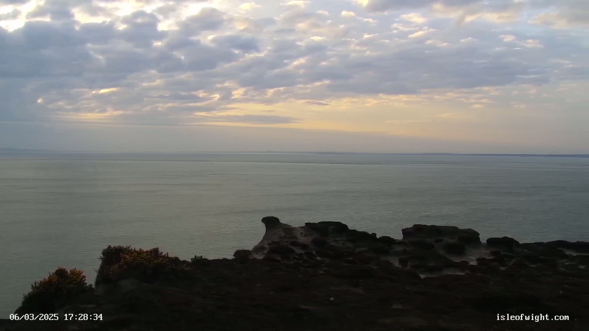 A rocky, dark coastline overlooks a calm sea under a partly cloudy, pastel sky at sunset.