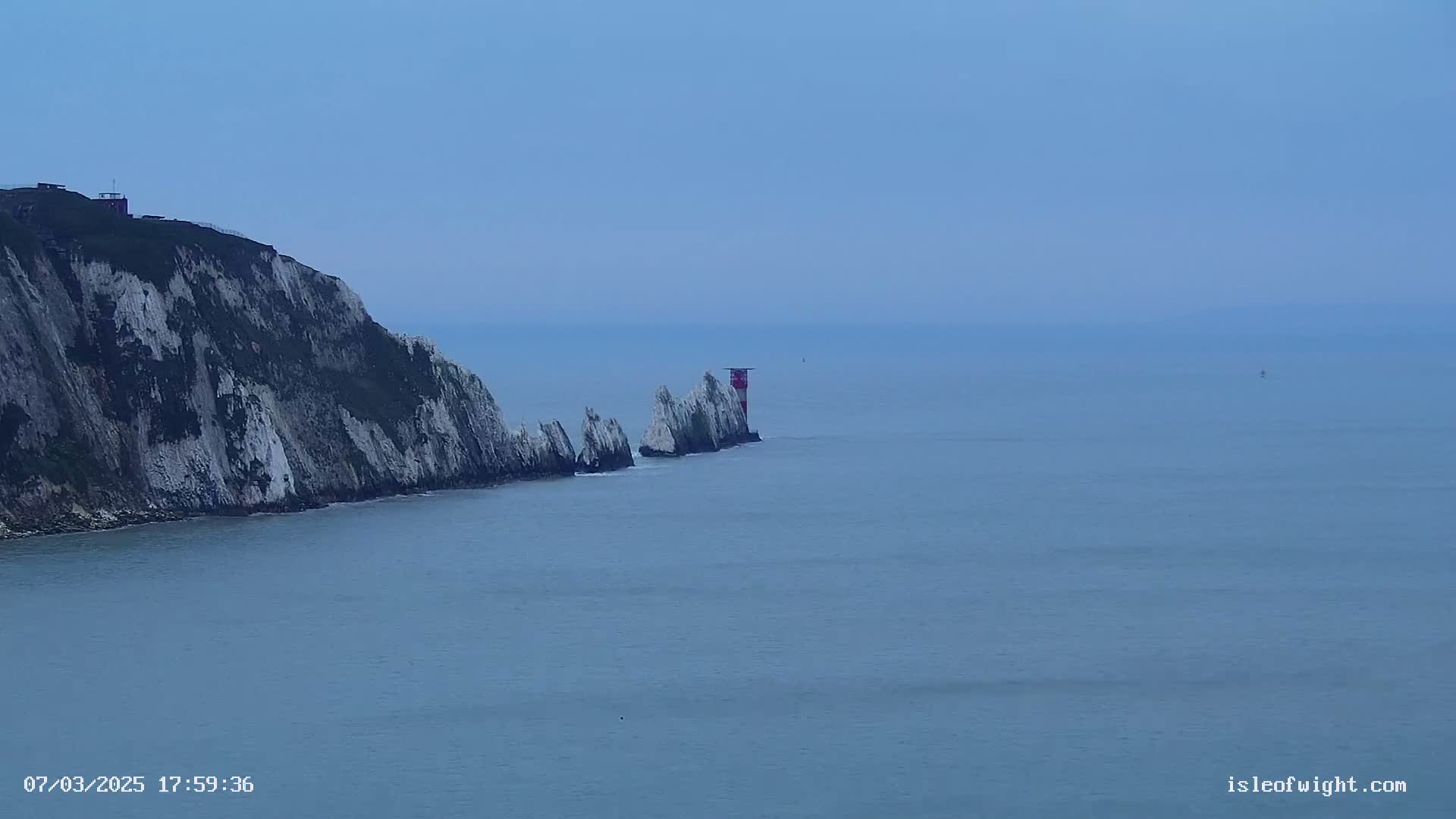 White cliffs meet a calm, blue sea where a small lighthouse stands atop a line of rocks.
