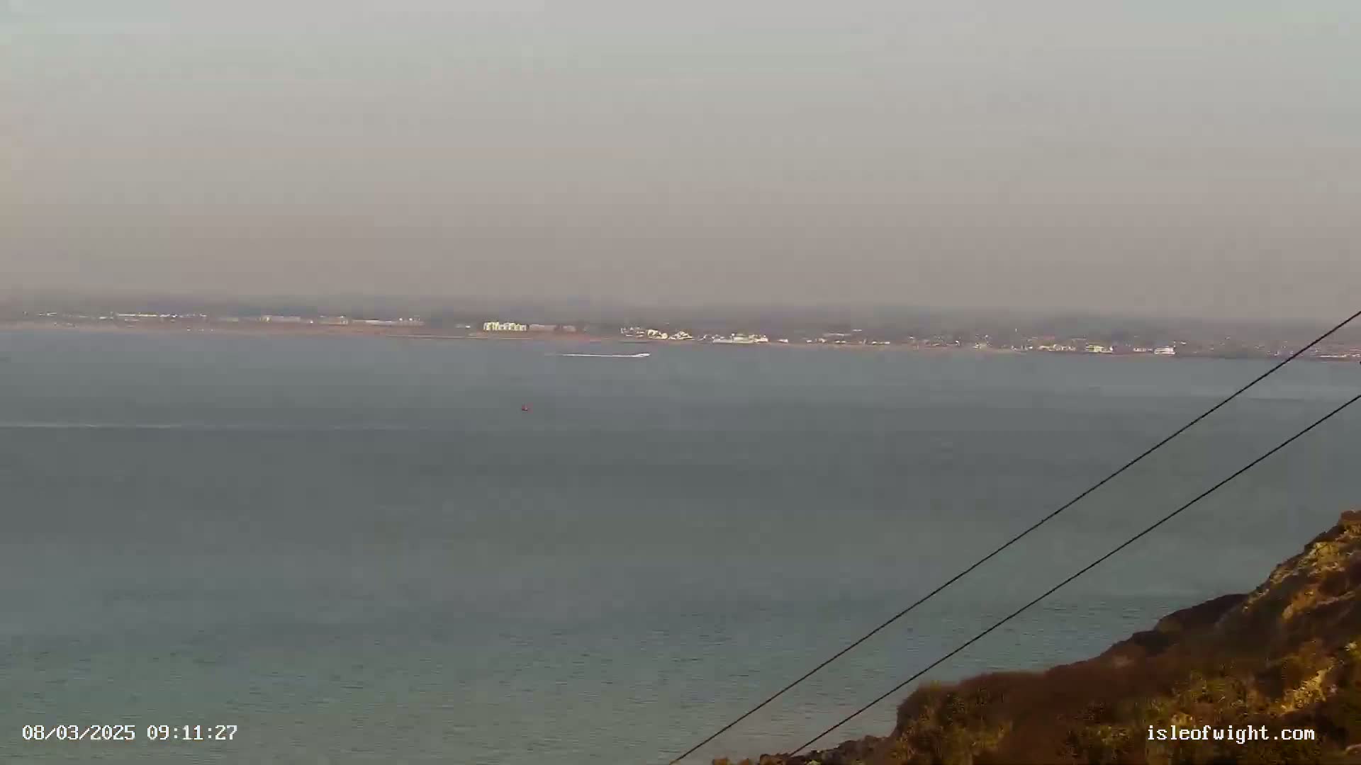 A hazy view of a calm body of water, with a distant shoreline showing buildings and a small boat visible on the water, seen from a high vantage point.