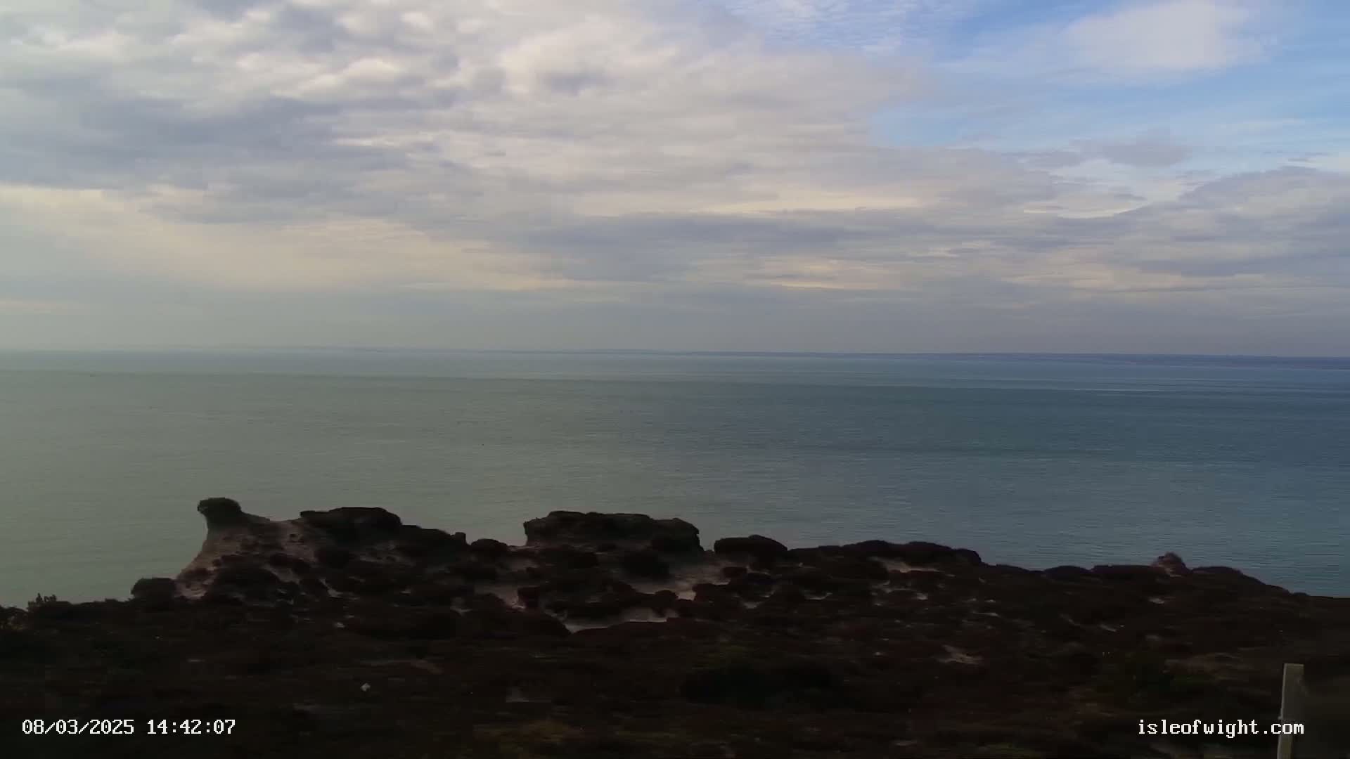 A rocky, dark-colored coastline overlooks a calm, blue-gray sea under a mostly cloudy sky.