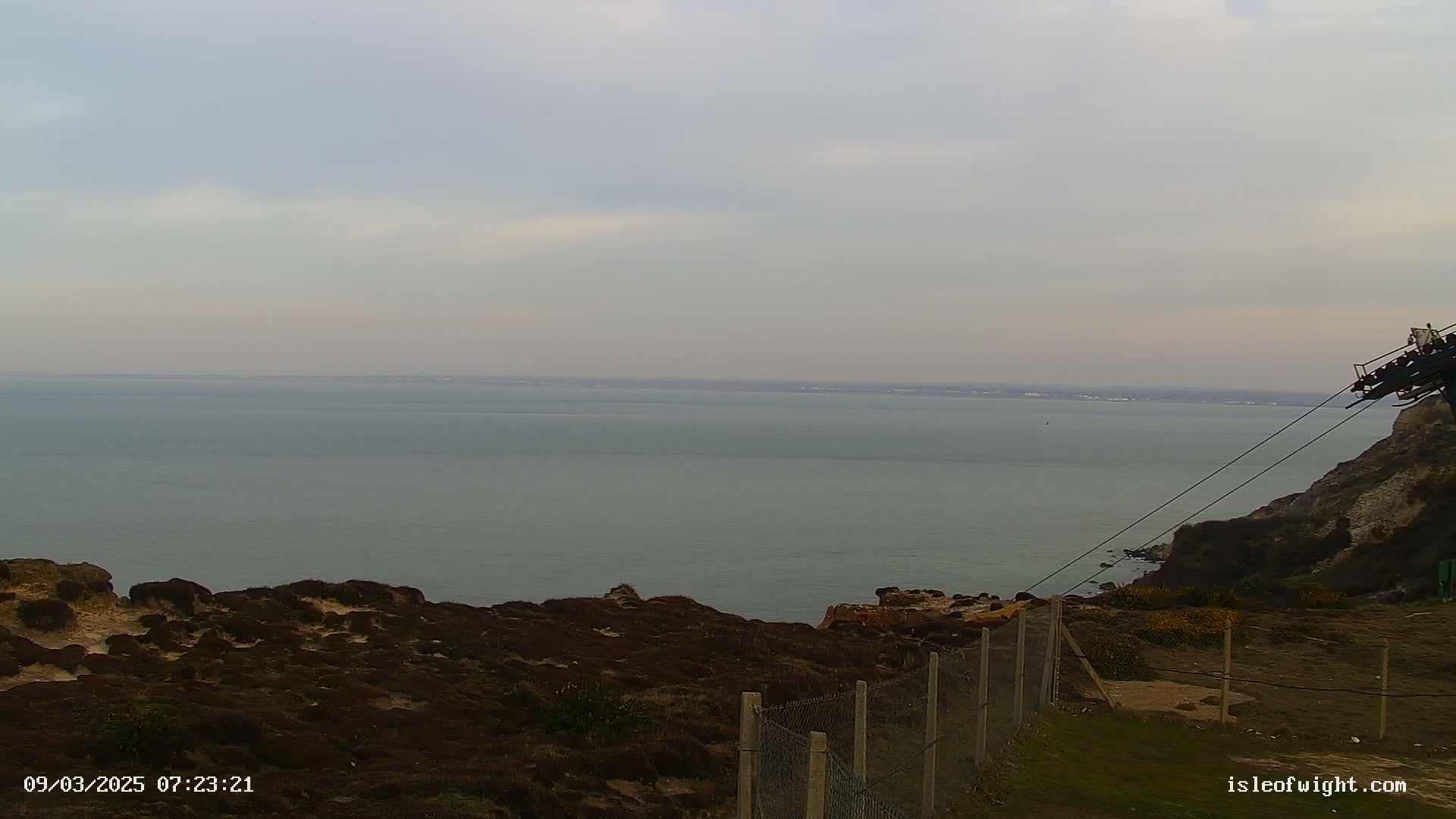 A calm, overcast day reveals a vast expanse of sea stretching to a distant shoreline, viewed from a cliff top with a chain-link fence and part of a cable car system visible.