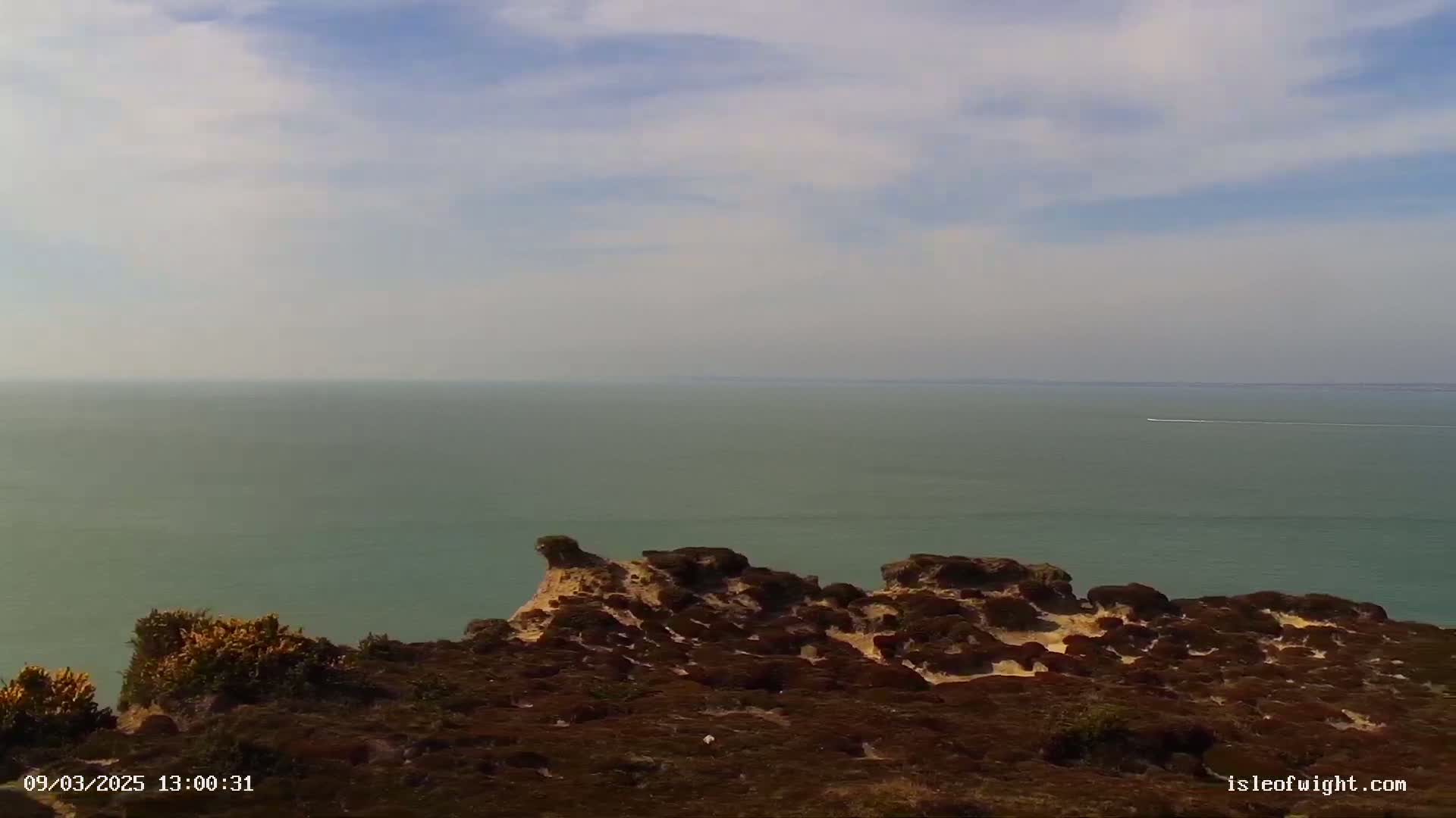 A tranquil expanse of sea under a partly cloudy sky is viewed from a high vantage point overlooking a rocky, shrub-covered coastline.