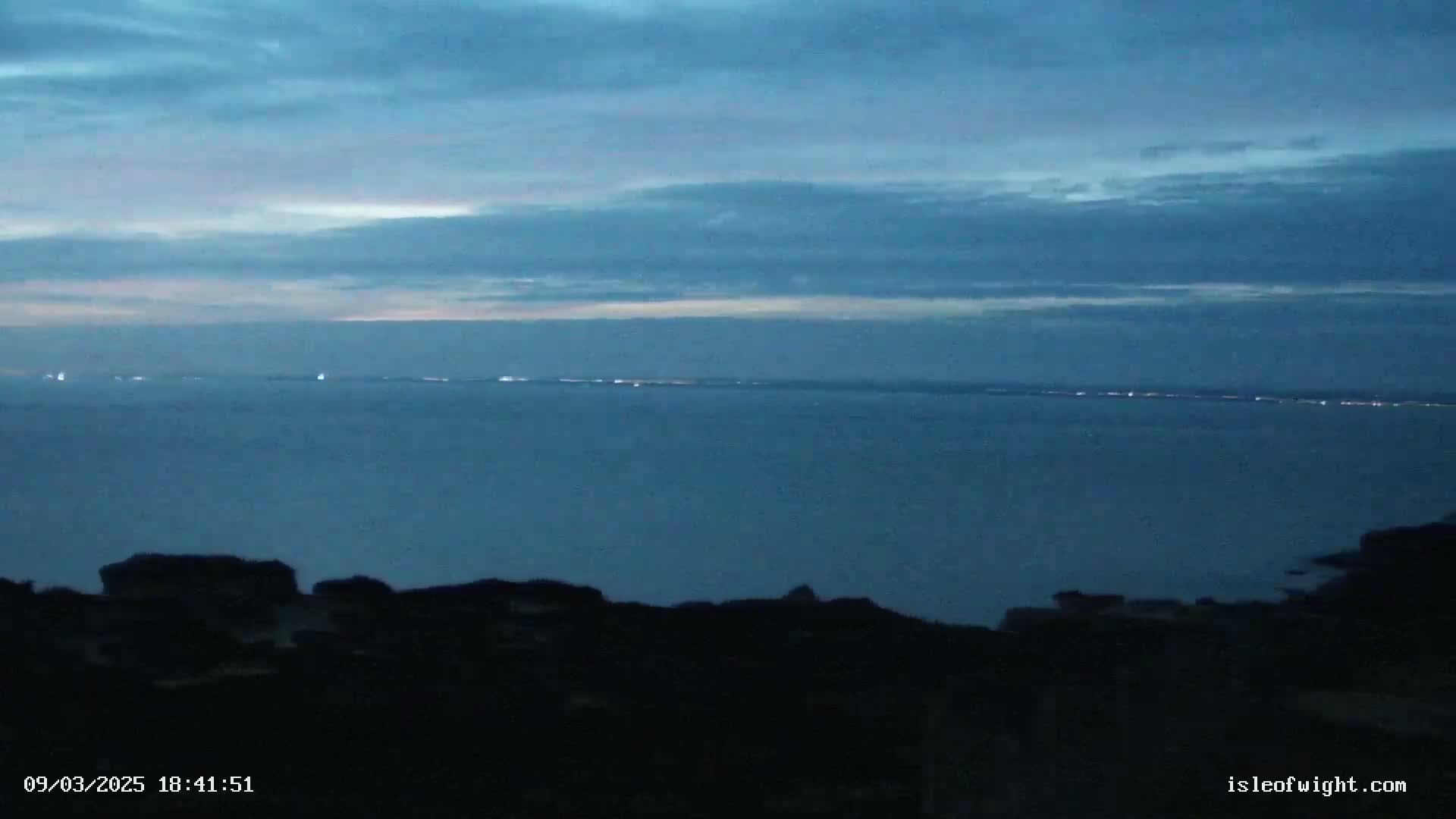 A dark, silhouetted coastal landscape at twilight shows a calm, dark-blue sea with a faint line of distant lights on the horizon under a mostly clear, dusky sky.