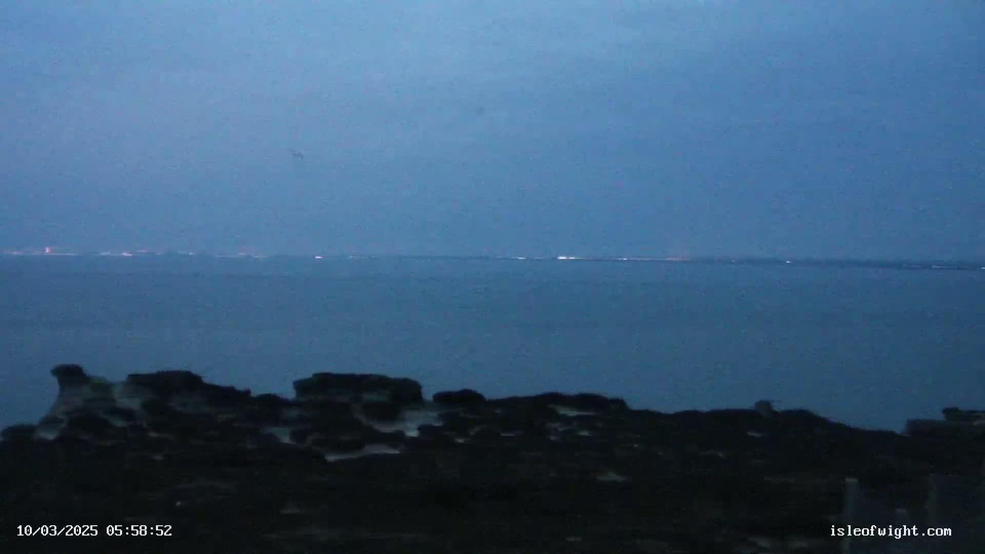 A dark, rocky shoreline overlooks a calm, blue-grey sea under a dusky sky at twilight.