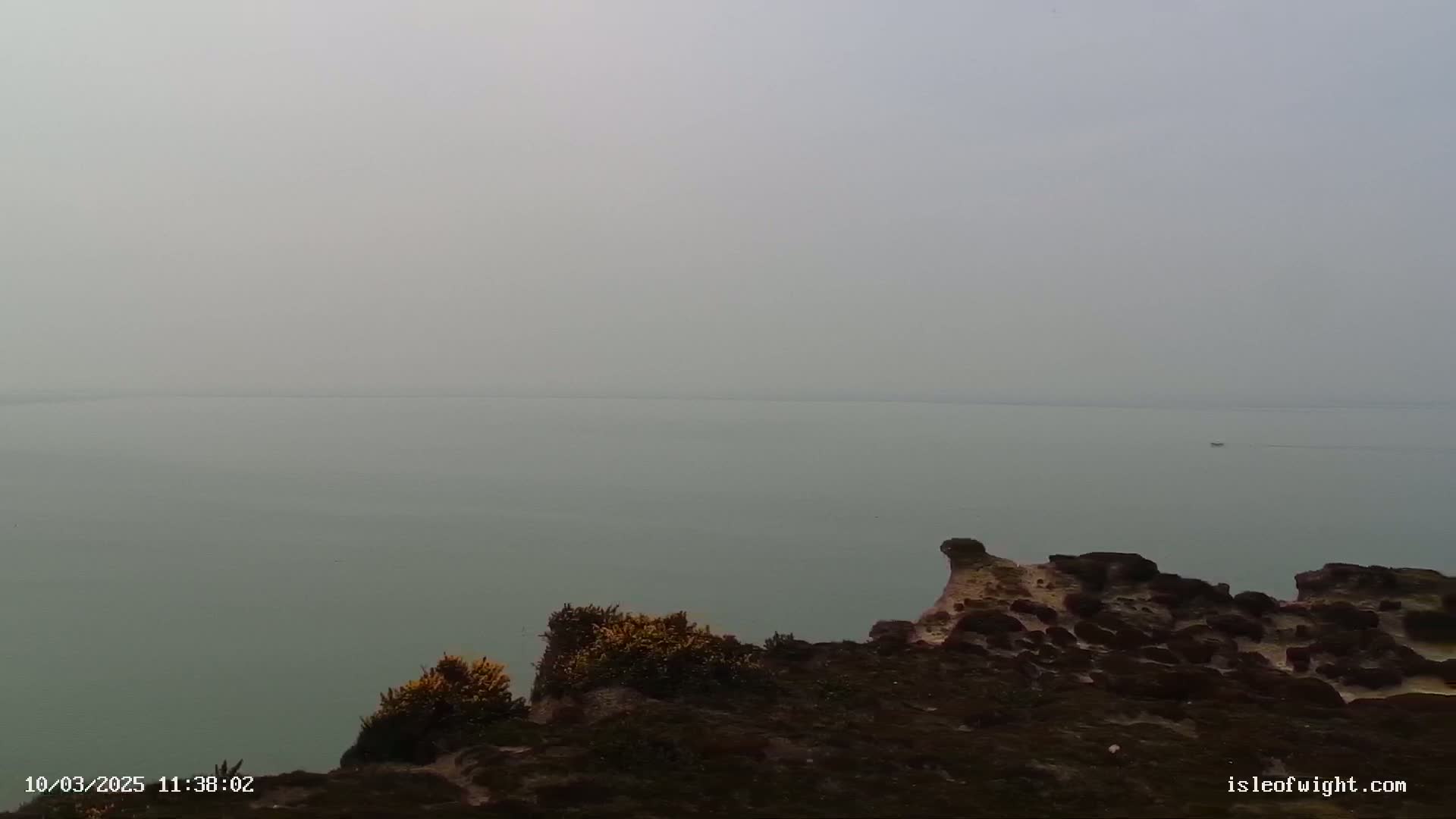 A hazy, calm body of water is visible from a rocky clifftop covered in low-lying vegetation.