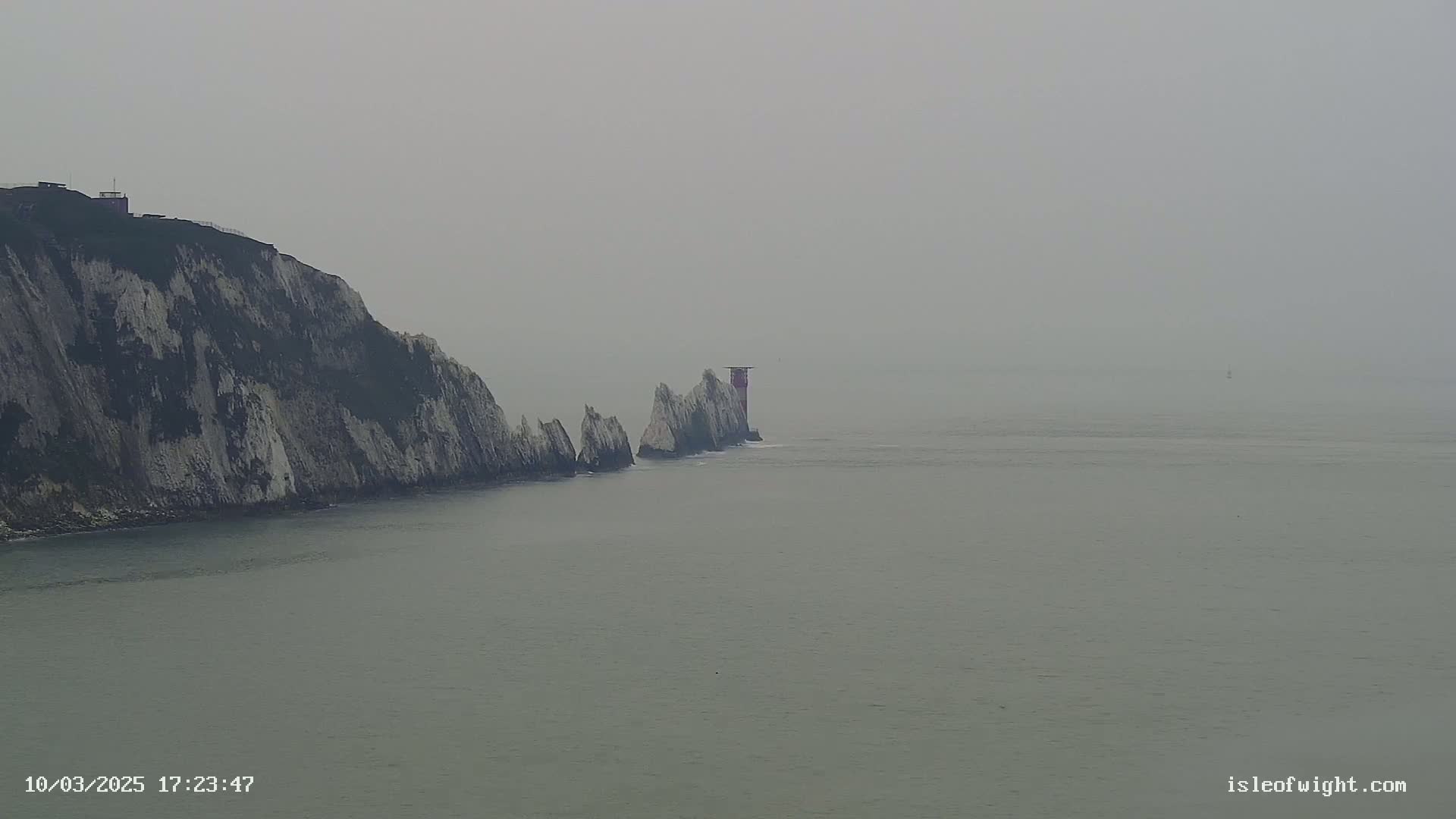 A lighthouse stands atop a series of white cliffs extending into a foggy sea.