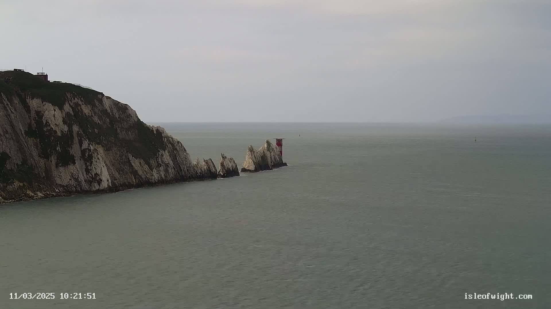 A white cliff faces a calm, gray sea where a series of rocky outcrops extend from the cliff toward a small red and white lighthouse.