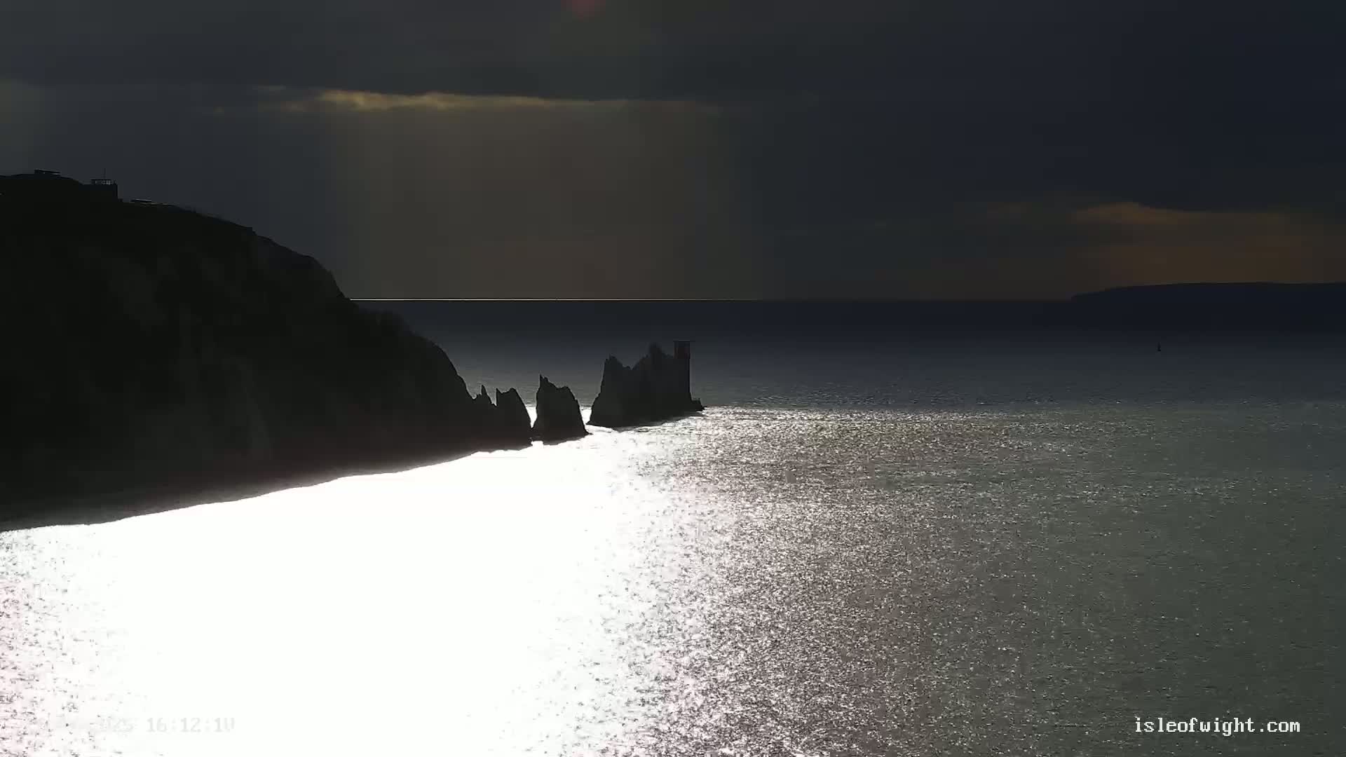Silhouetted cliffs and rock formations jut into a sparkling sea under a dark, overcast sky.