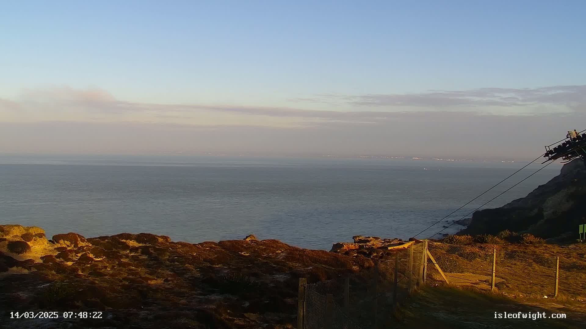 A calm, hazy ocean stretches to the horizon from a rocky clifftop, partially enclosed by a wire fence.