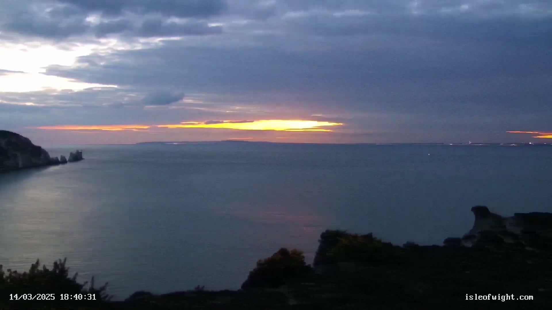 A sunset over a calm sea with a rocky coastline, under a mostly cloudy sky.