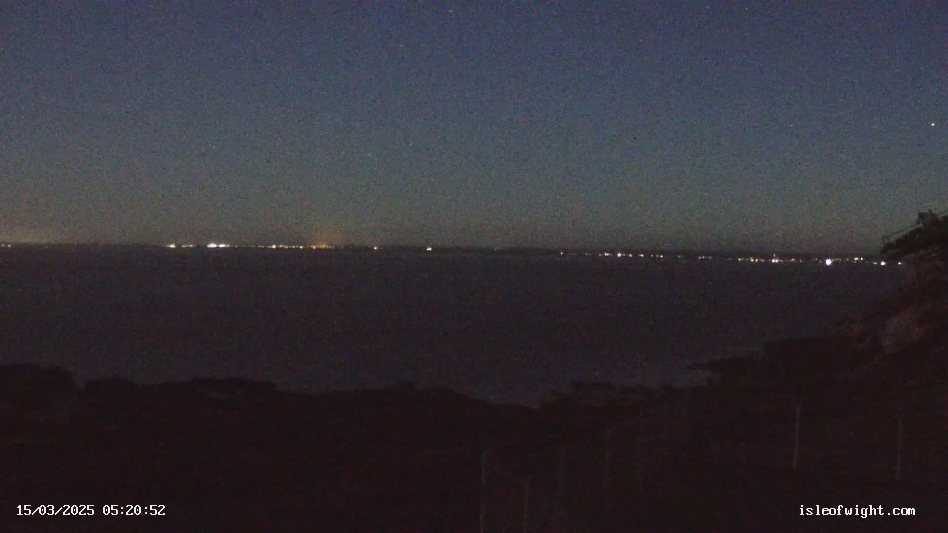 A dark, nighttime scene shows a distant line of lights across a body of water under a clear, starry sky.