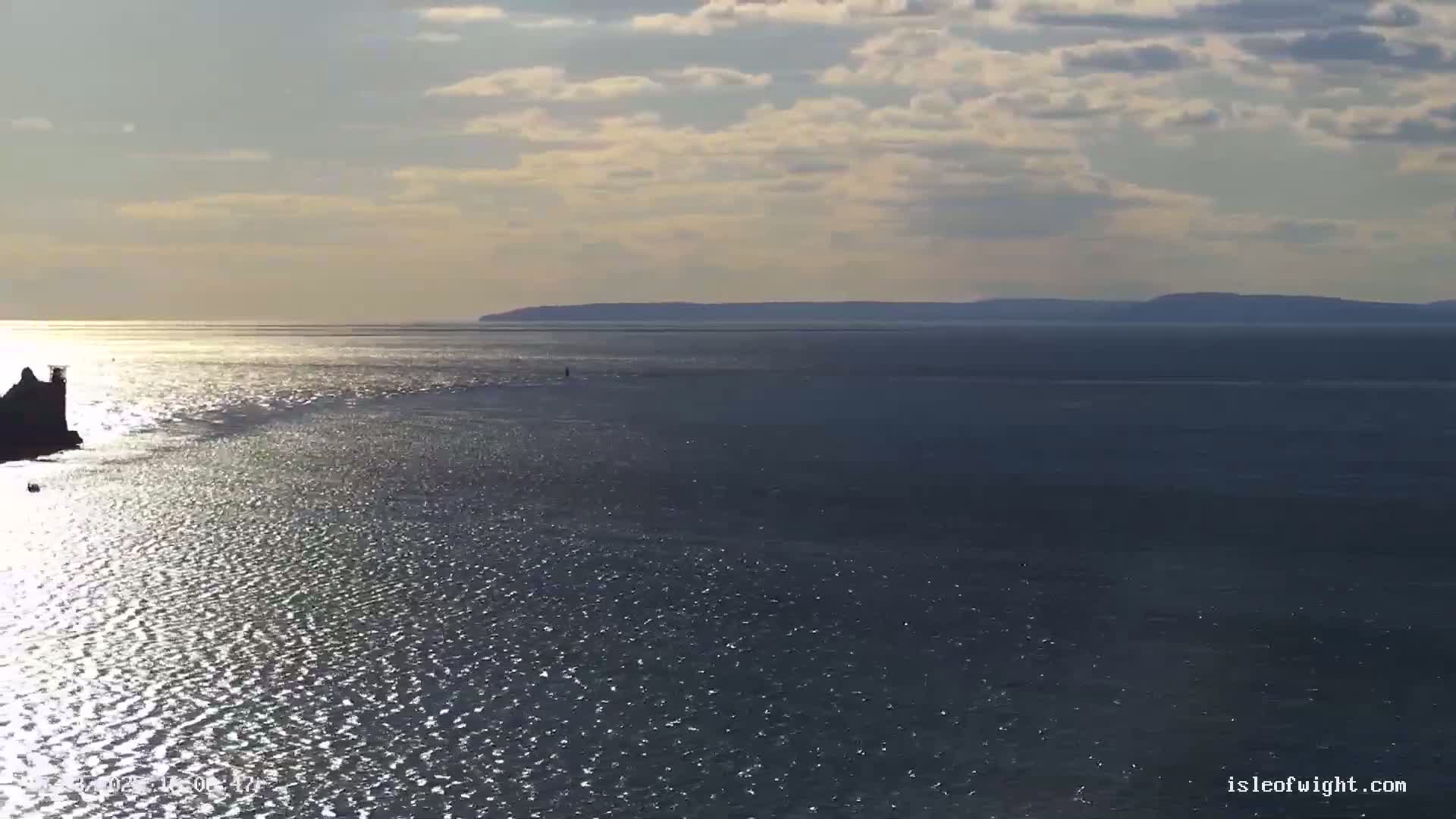 A partly cloudy day shows a wide expanse of glittering water, with a distant landmass visible on the horizon and a dark rocky outcrop in the foreground.