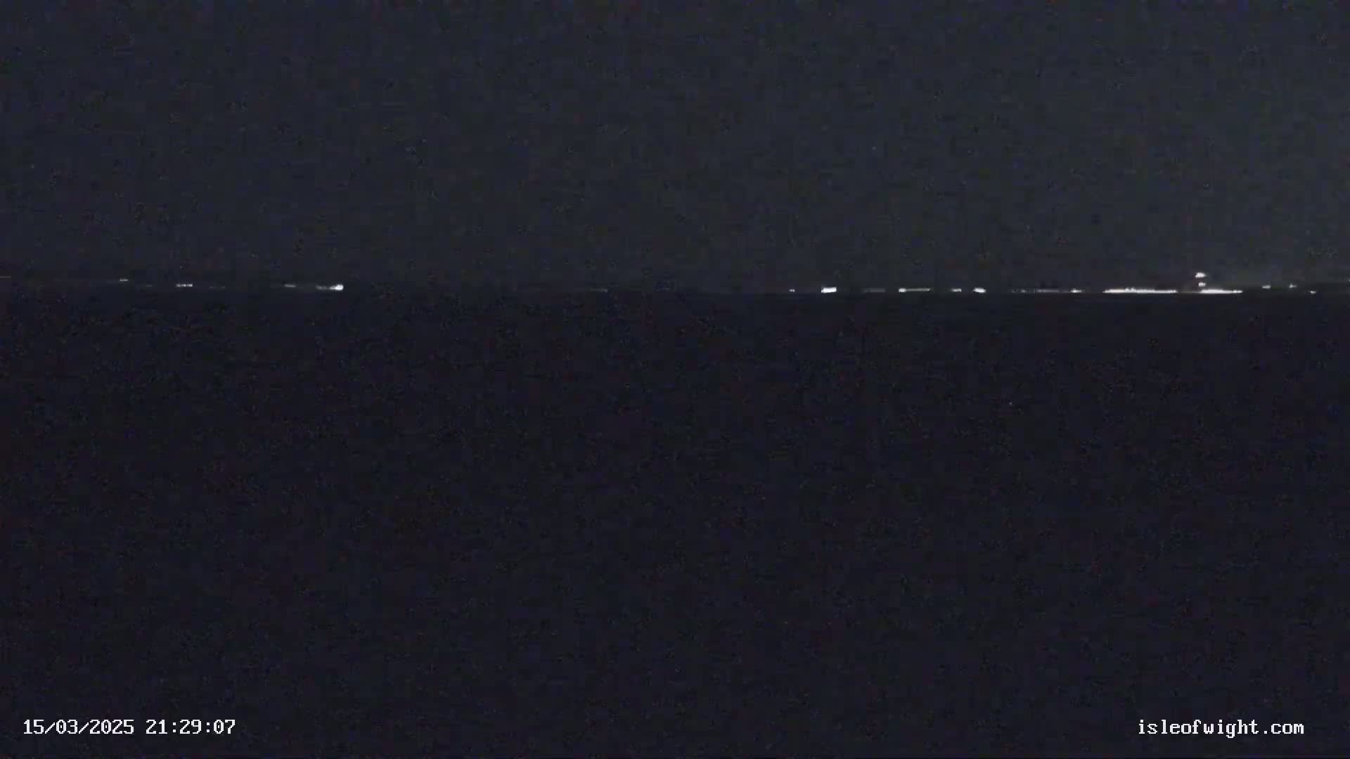 A dark nighttime scene shows a faint line of lights on the horizon over a dark body of water.