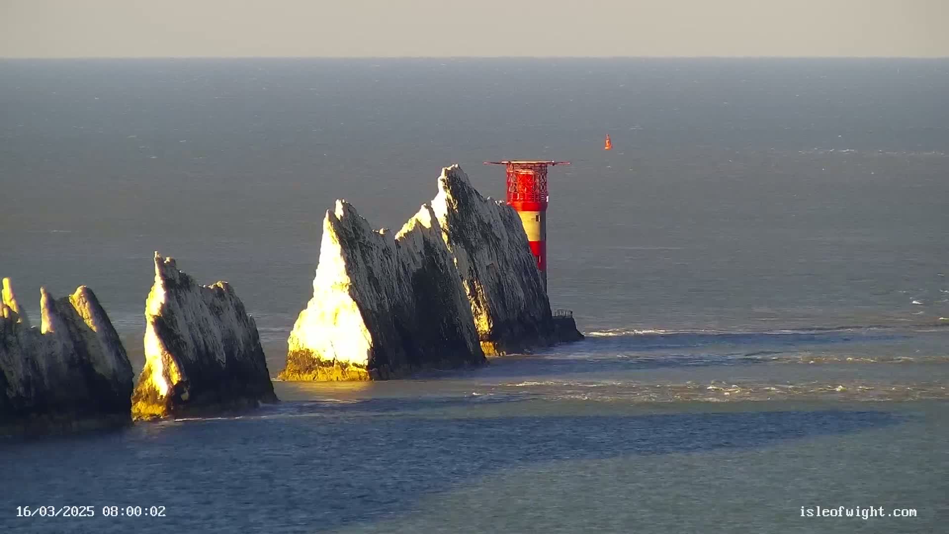 A red and white lighthouse stands atop a cluster of jagged white cliffs extending into a calm, sunlit sea.