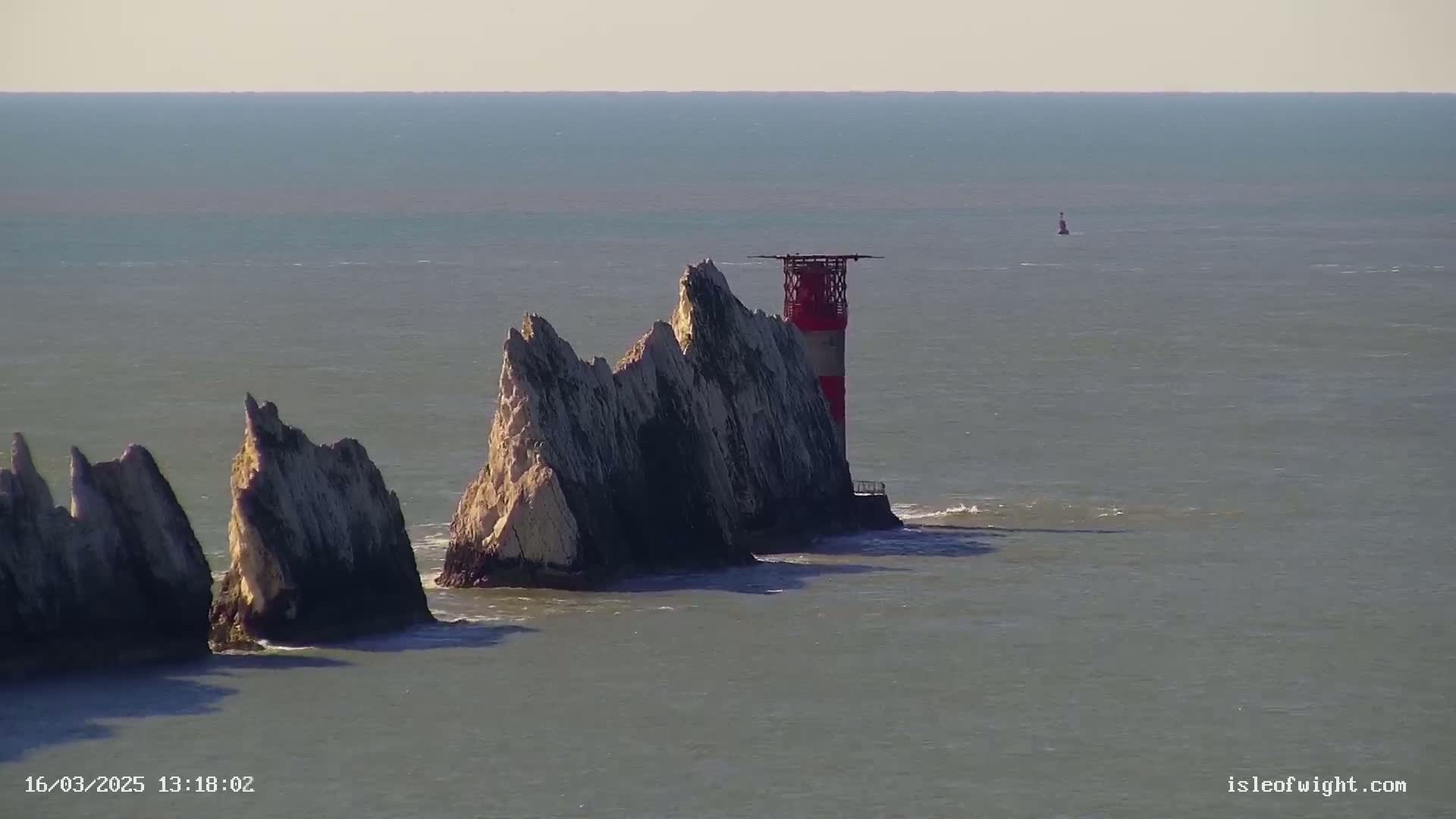 A red and white lighthouse stands atop a cluster of jagged, light-colored rocks extending into a calm, gray-blue sea under a clear sky.