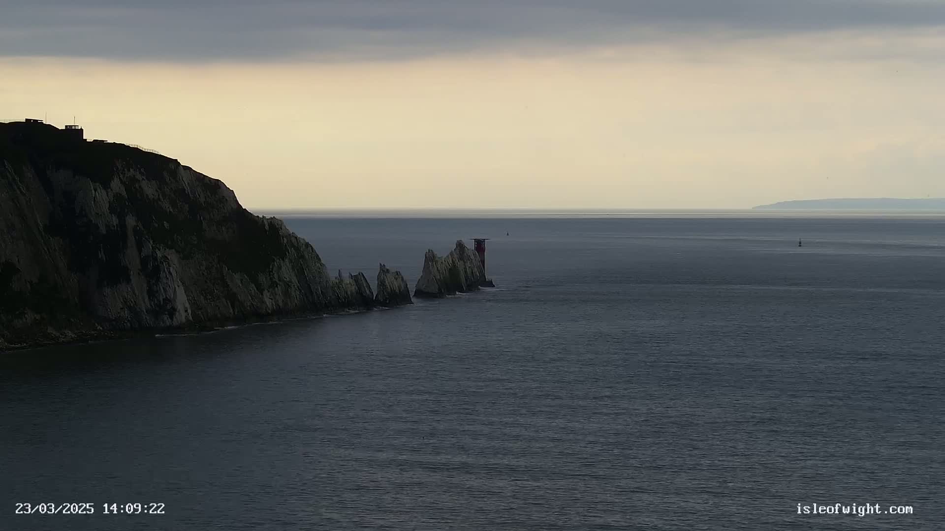 A rocky cliff overlooks a calm sea with a line of smaller rock formations and a small lighthouse extending into the water under a cloudy sky.