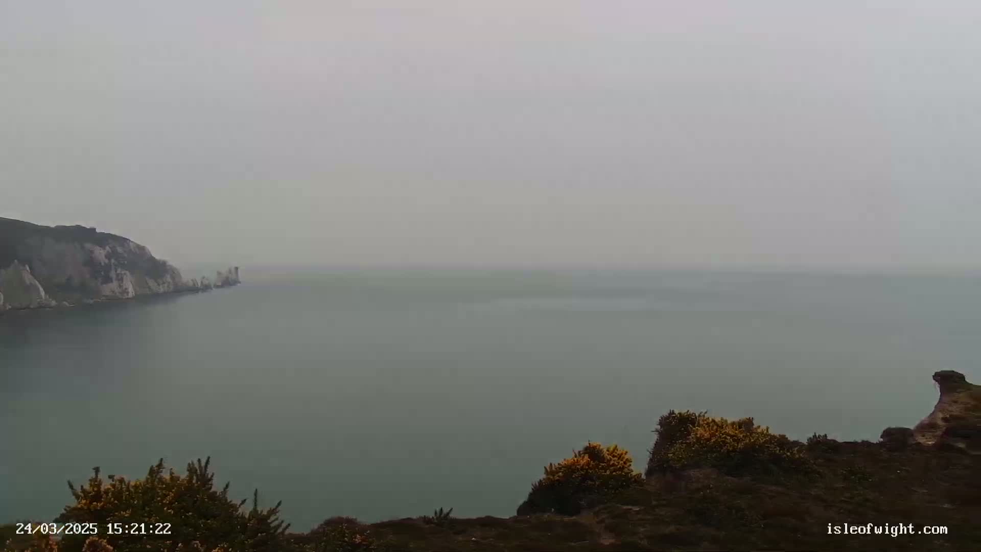 A hazy view of a calm sea stretches to the horizon from a clifftop, where low-lying vegetation is visible.