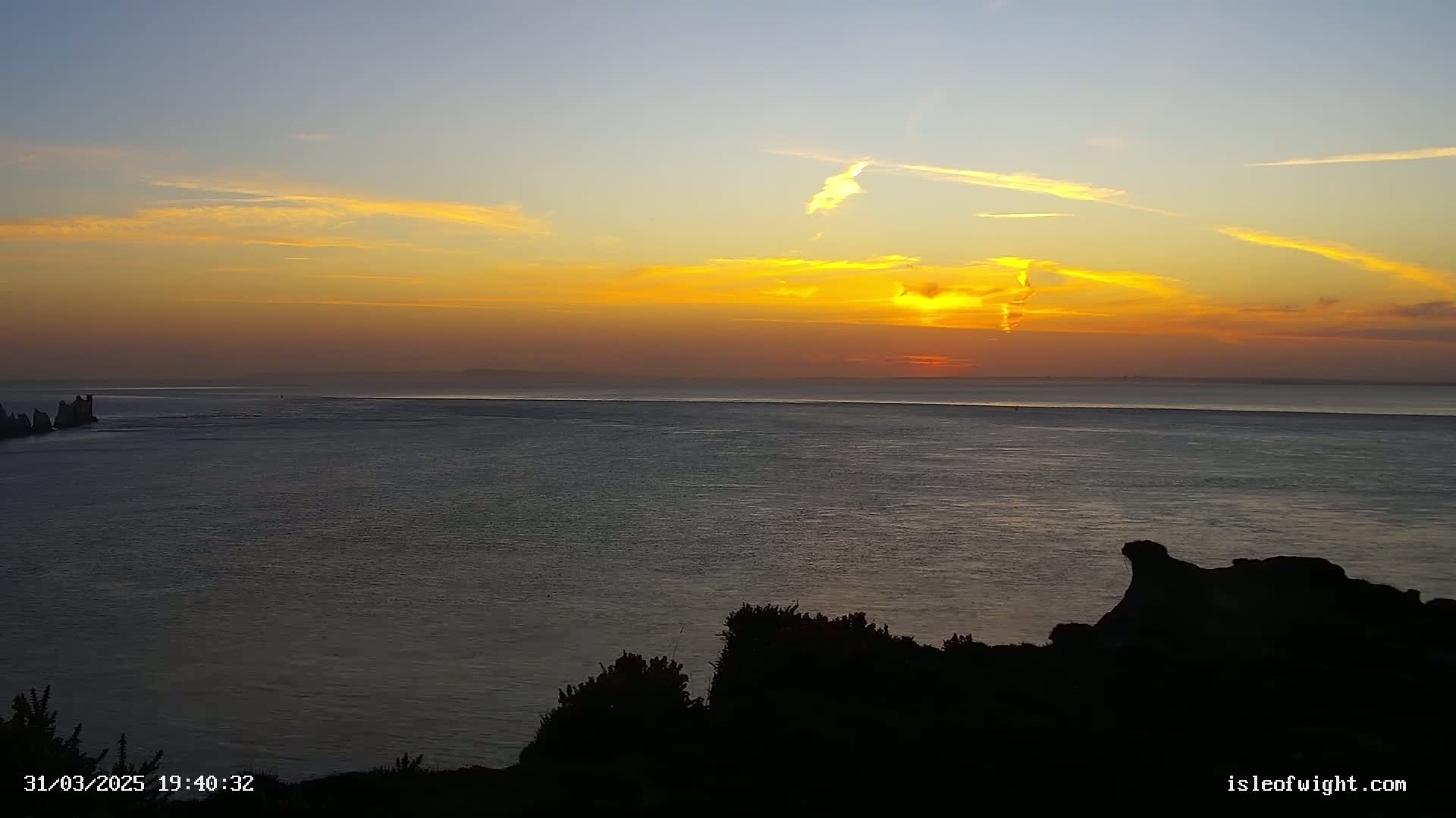 A sunset over a calm sea with a distant coastline and silhouetted rocks in the foreground.