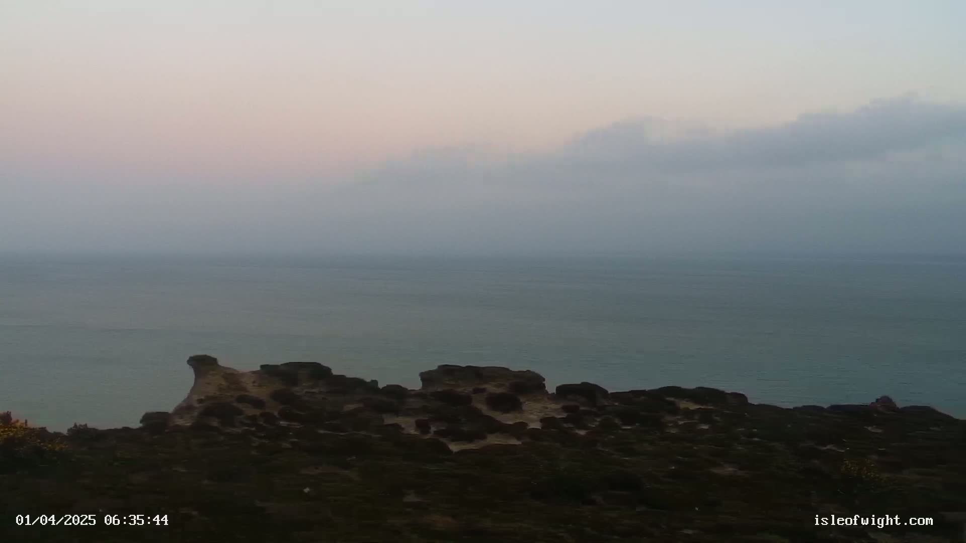 A hazy, pale sky overlooks a calm sea and a rocky, shrub-covered coastline.
