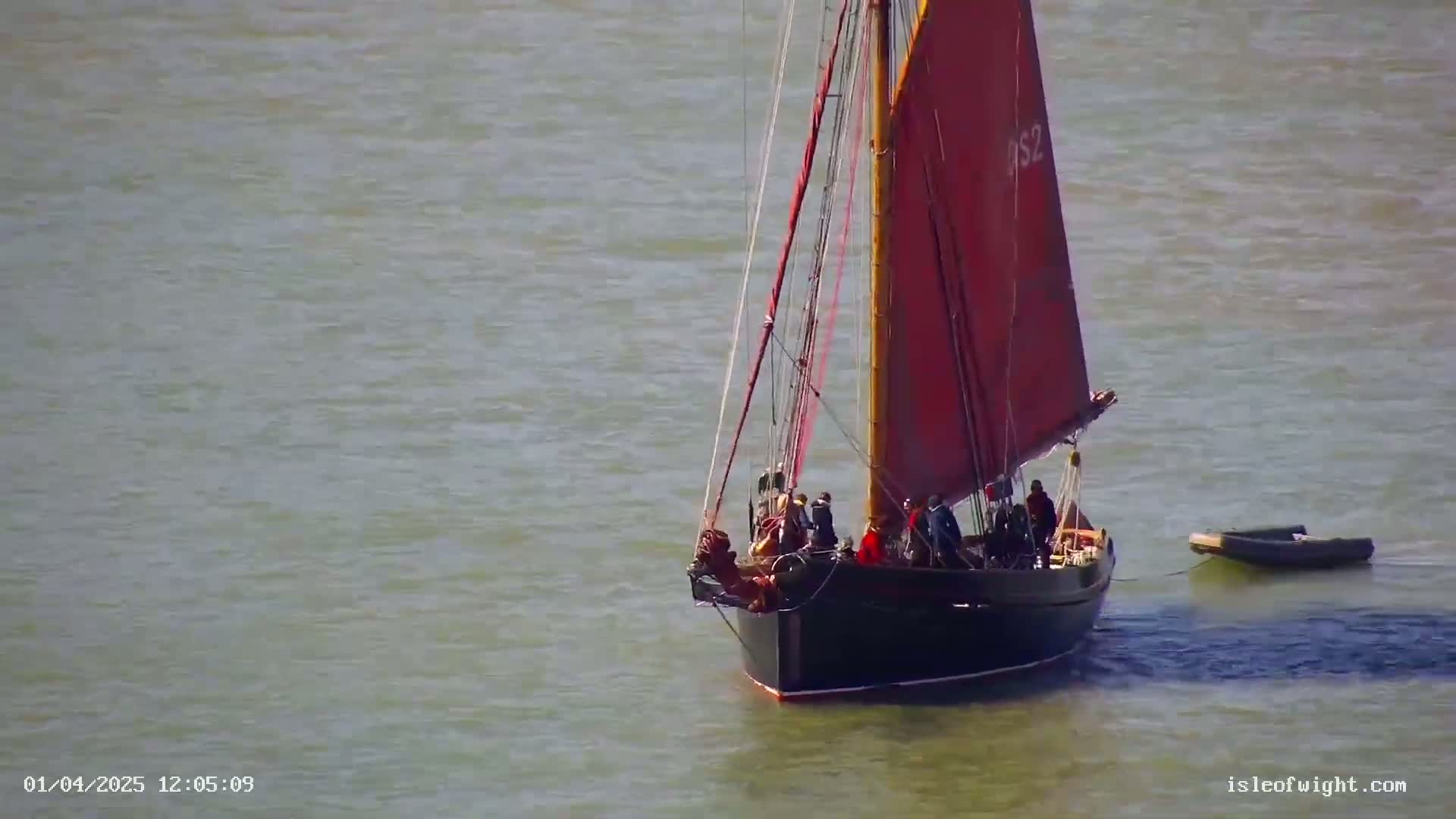 A black sailboat with a red sail and several people aboard floats on calm, gray water, with a small inflatable boat trailing behind it.