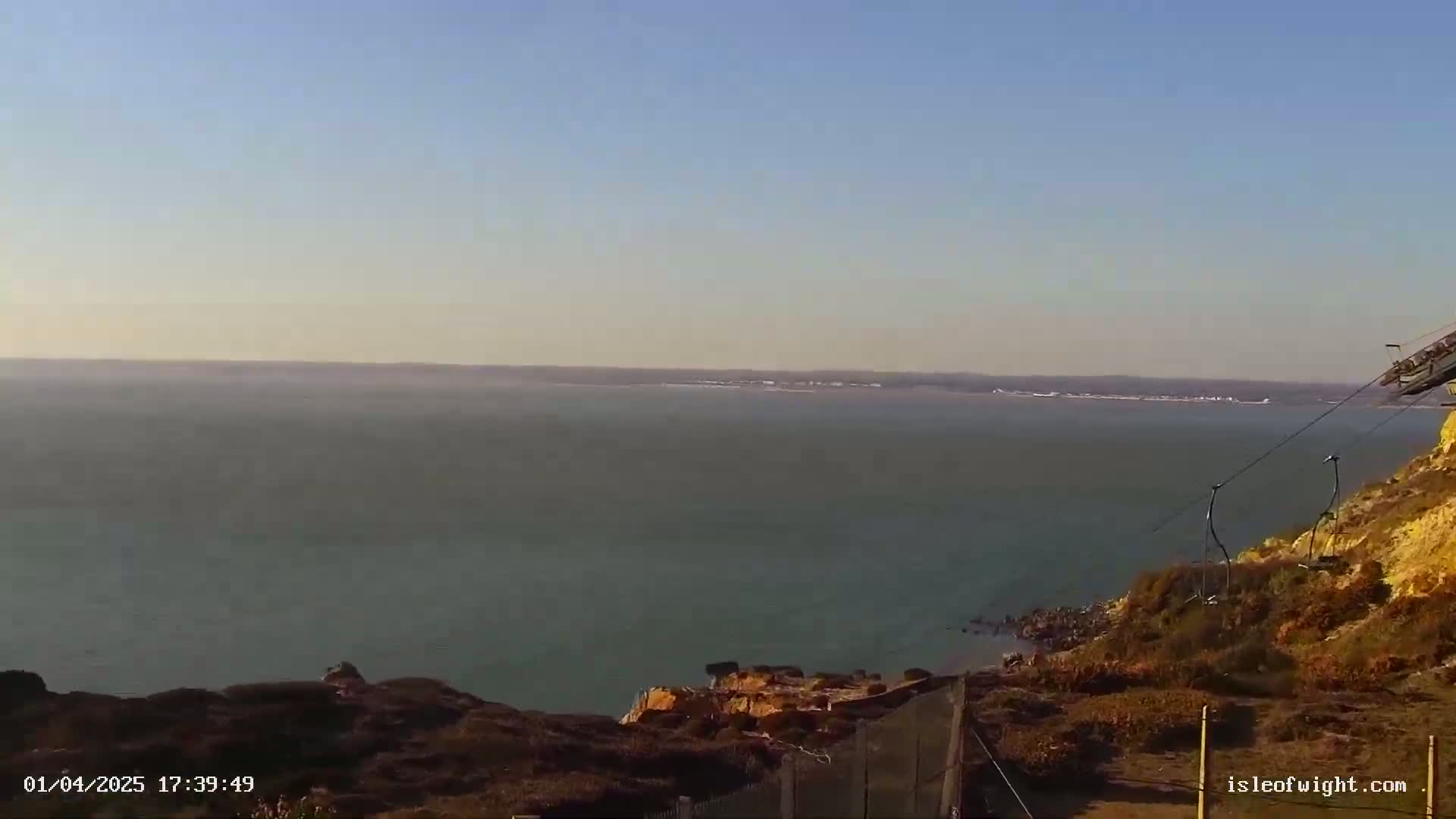 A calm body of water stretches to a hazy horizon under a clear sky, viewed from a cliff top with sparse vegetation and a chairlift partially visible.