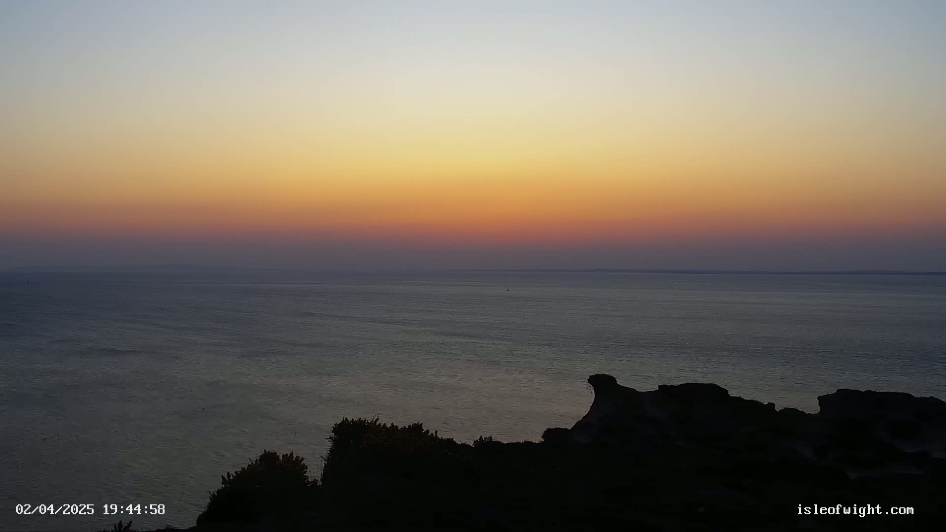 A sunset over a calm ocean is viewed from a rocky, silhouetted coastal vantage point.