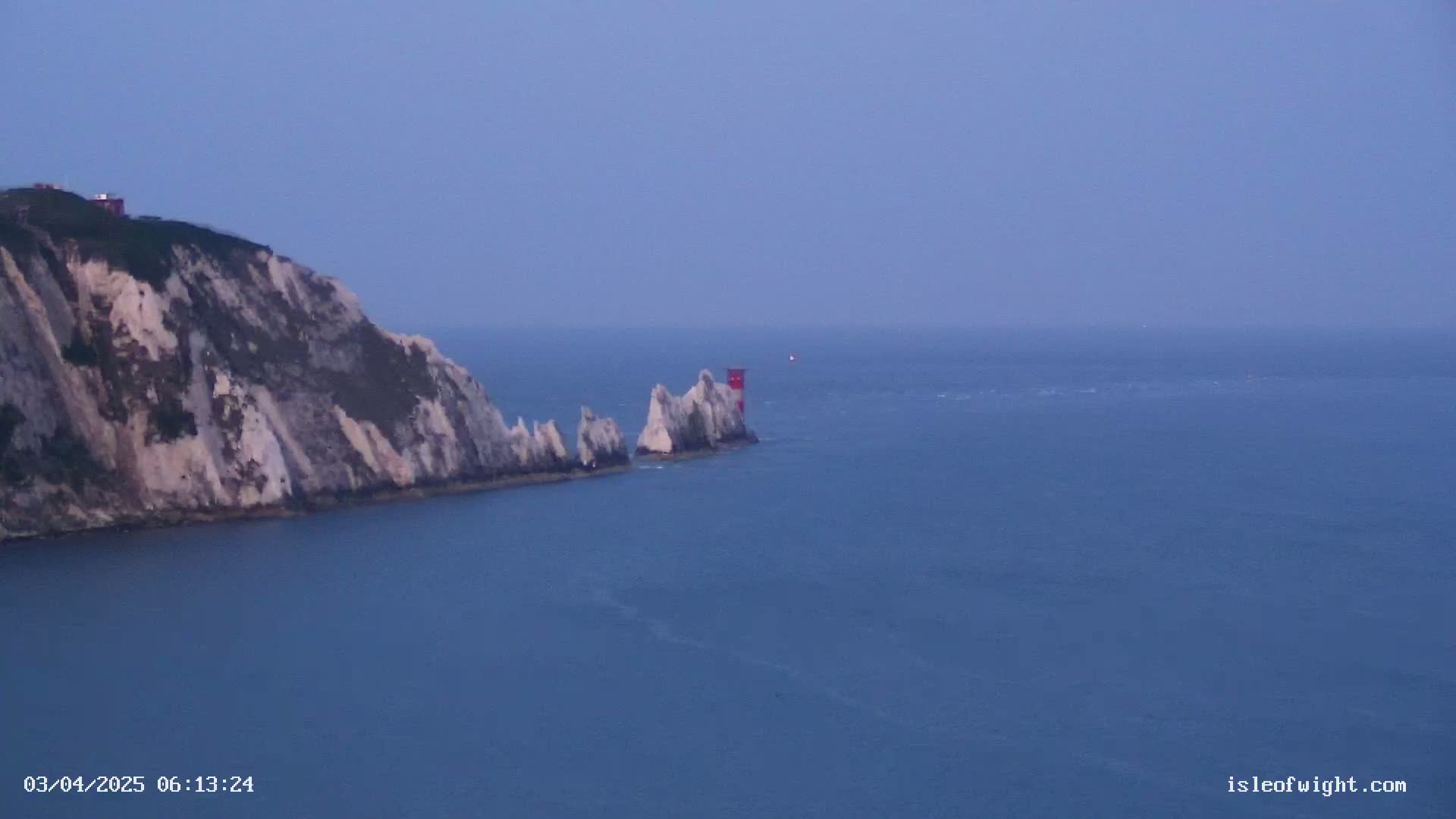 A white cliff overlooks a calm, blue sea where a small, red and white lighthouse stands near a cluster of white rocks.