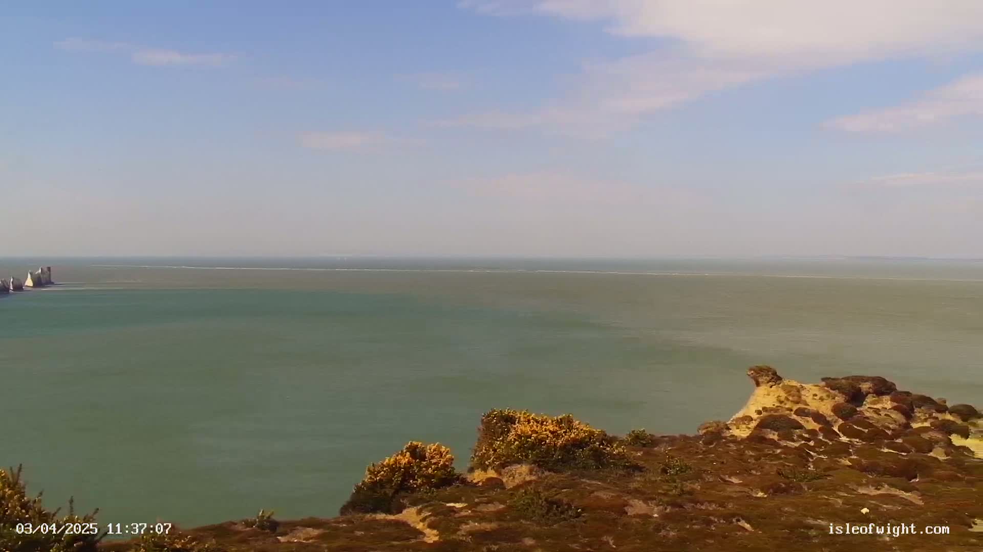 A view from a clifftop shows a calm, greenish-grey sea extending to a distant horizon under a mostly clear, light blue sky.