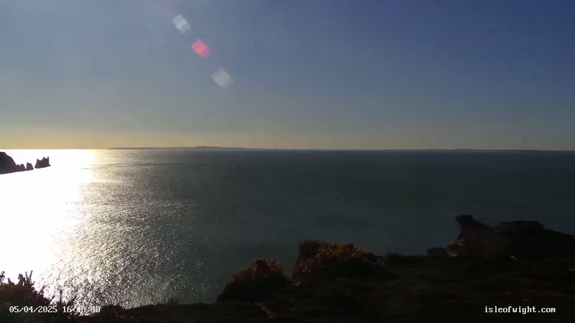 A sunlit expanse of ocean stretches to a distant shoreline under a clear, bright sky, with dark rocky land and vegetation in the foreground.