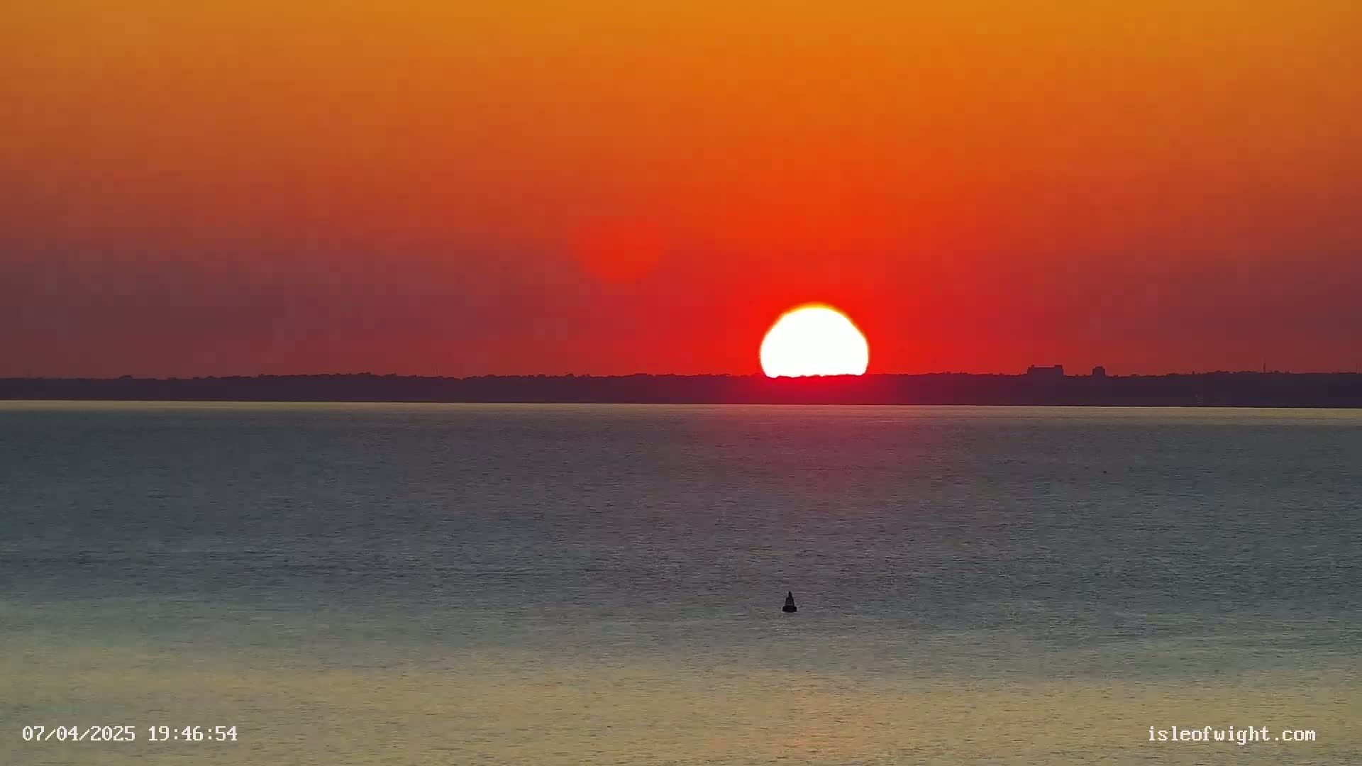 A large, bright sun sets over a calm body of water and a dark, distant shoreline under an orange and red sky.